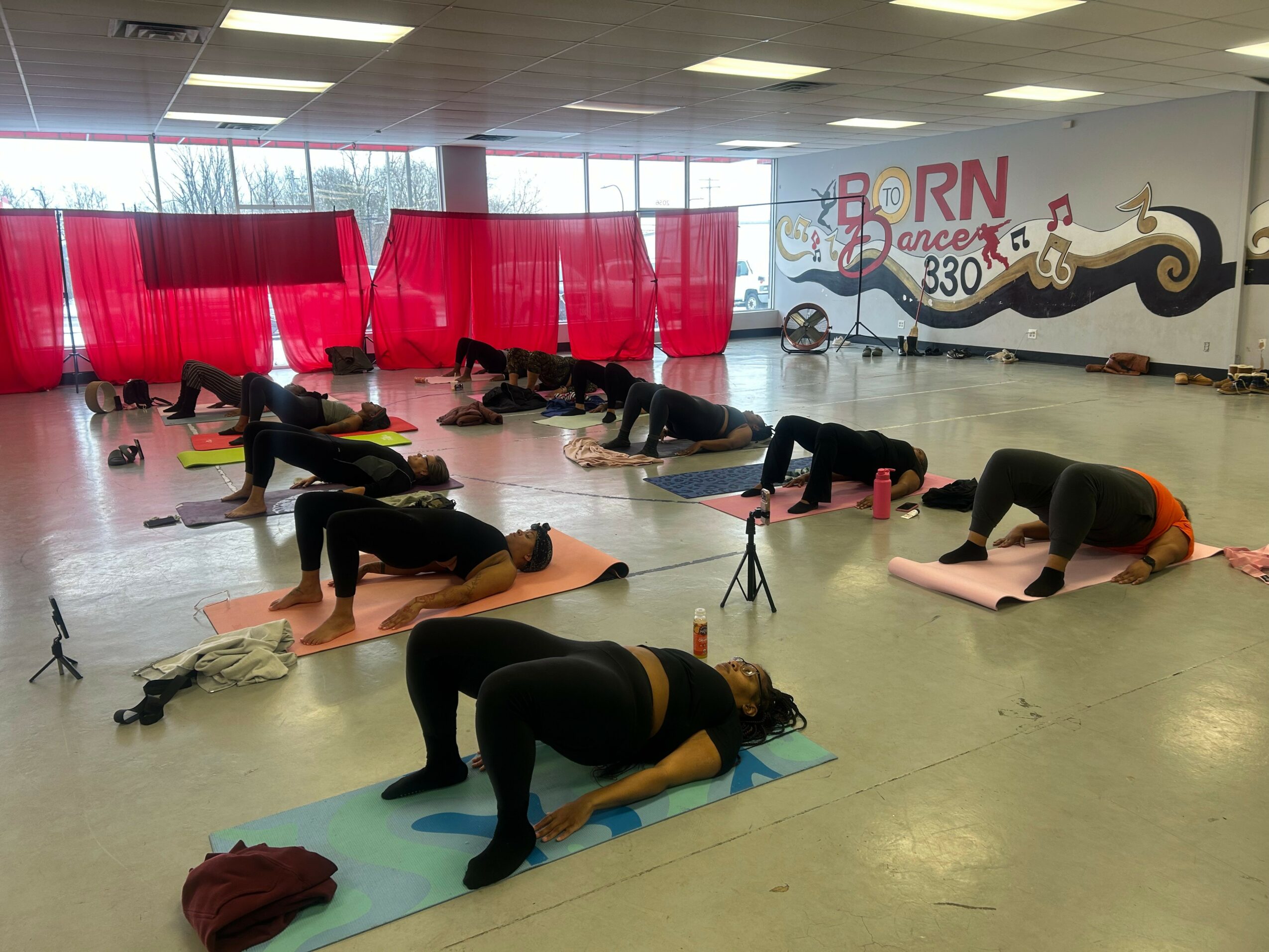 People practicing yoga on mats in a studio with red curtains and a mural on the wall, some in downward dog pose.