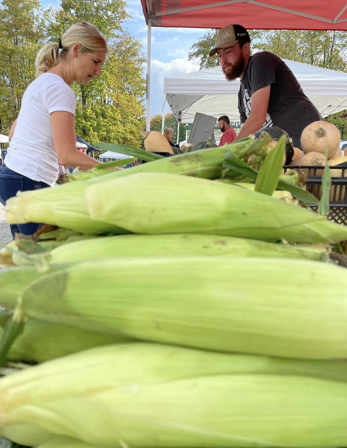 Two people at a market stall with corn and vegetables, outdoors with trees and tents in the background.