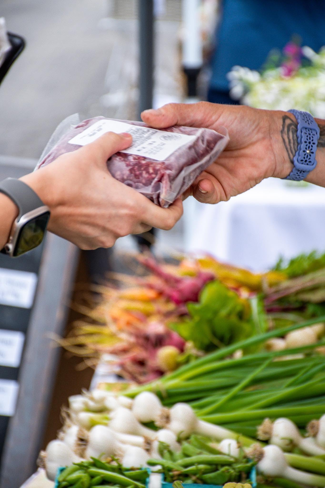 Person handing over a packaged item to another person at an outdoor market stall with flowers and vegetables.