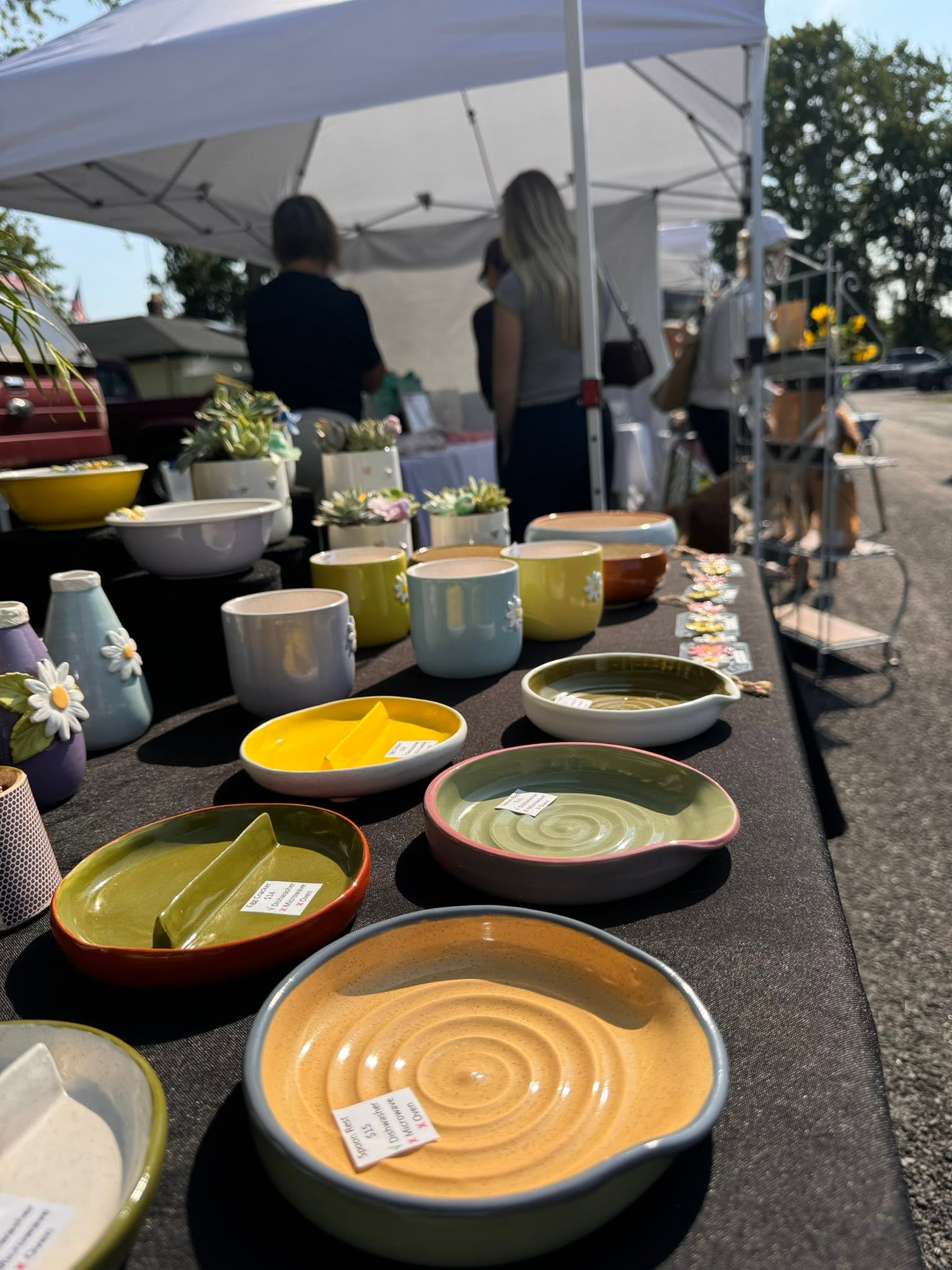 Display of colorful ceramic bowls and plates outdoors under a tent, with people browsing in the background.