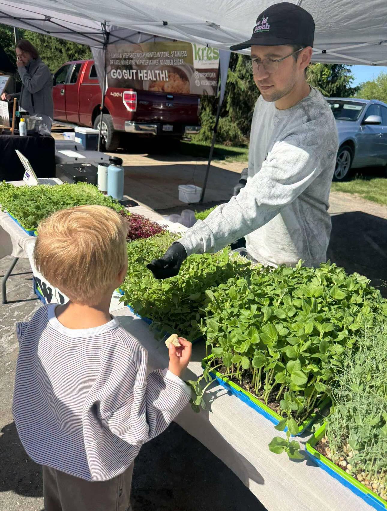 Young man wearing glasses and a cap sells fresh herbs at an outdoor market stall to a child with red hair, under a tent.