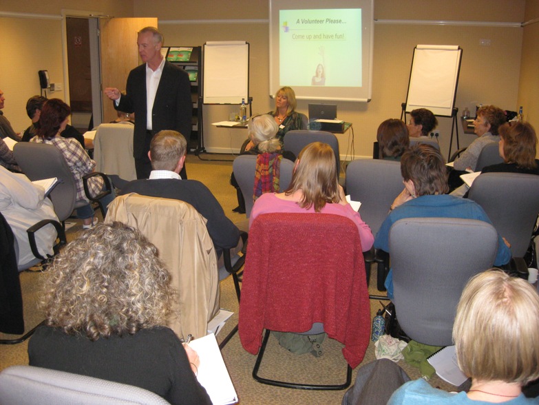 A man in a suit presents to a seated audience in a conference room with a projector screen and whiteboards.