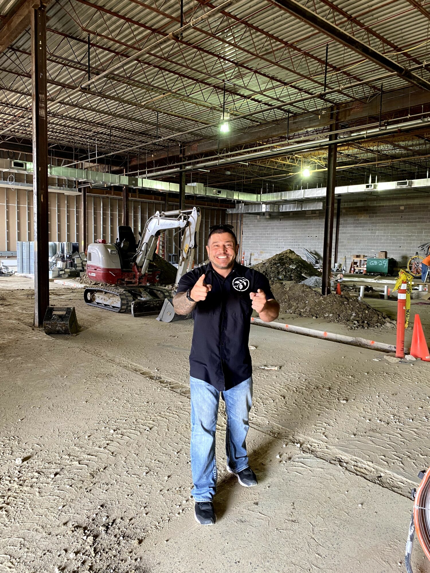 Man standing in an indoor construction site, smiling and giving a thumbs-up, with construction equipment and orange cones in background.
