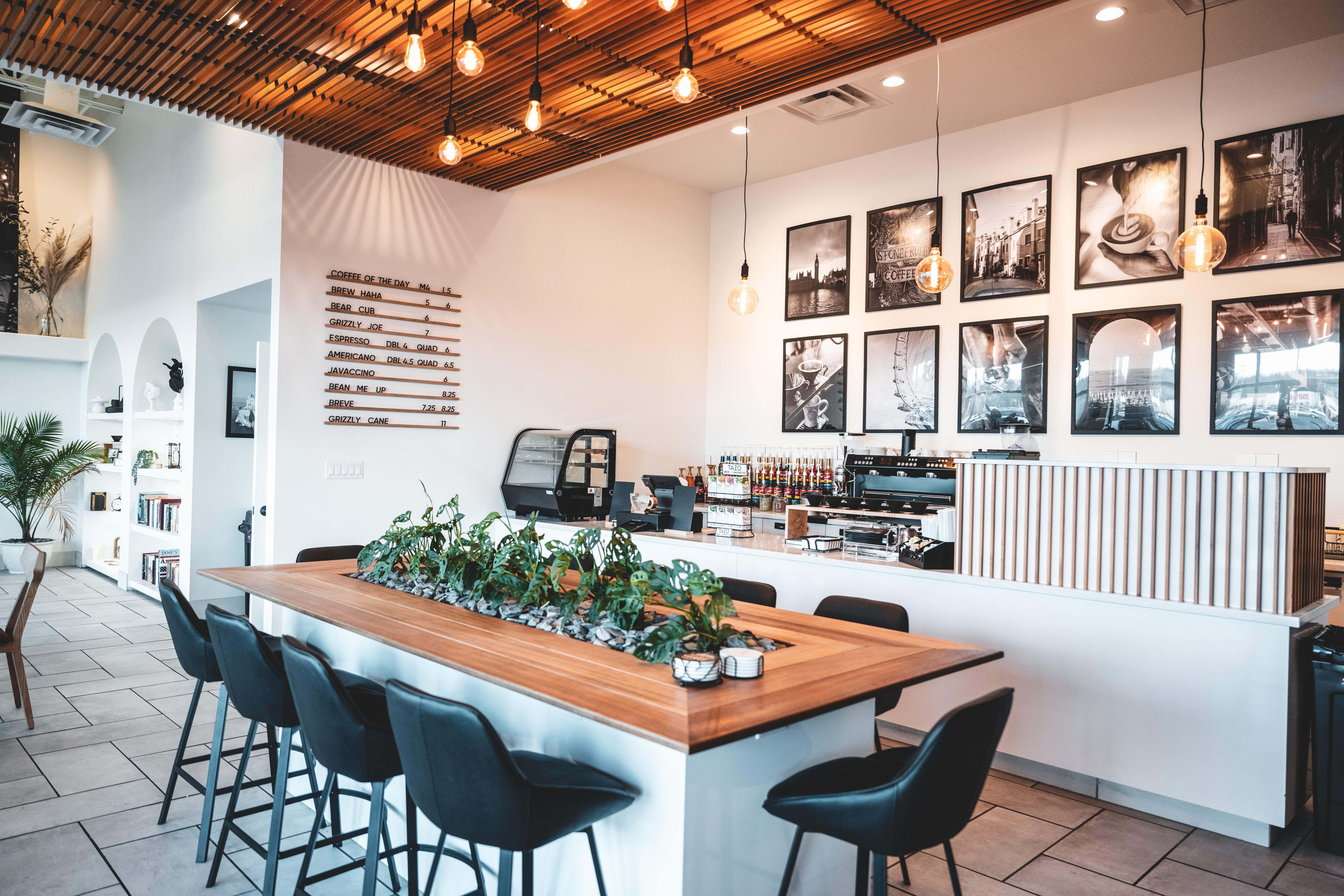 Modern cafe interior with a long wooden table, black chairs, framed photos on the wall, and hanging pendant lights.