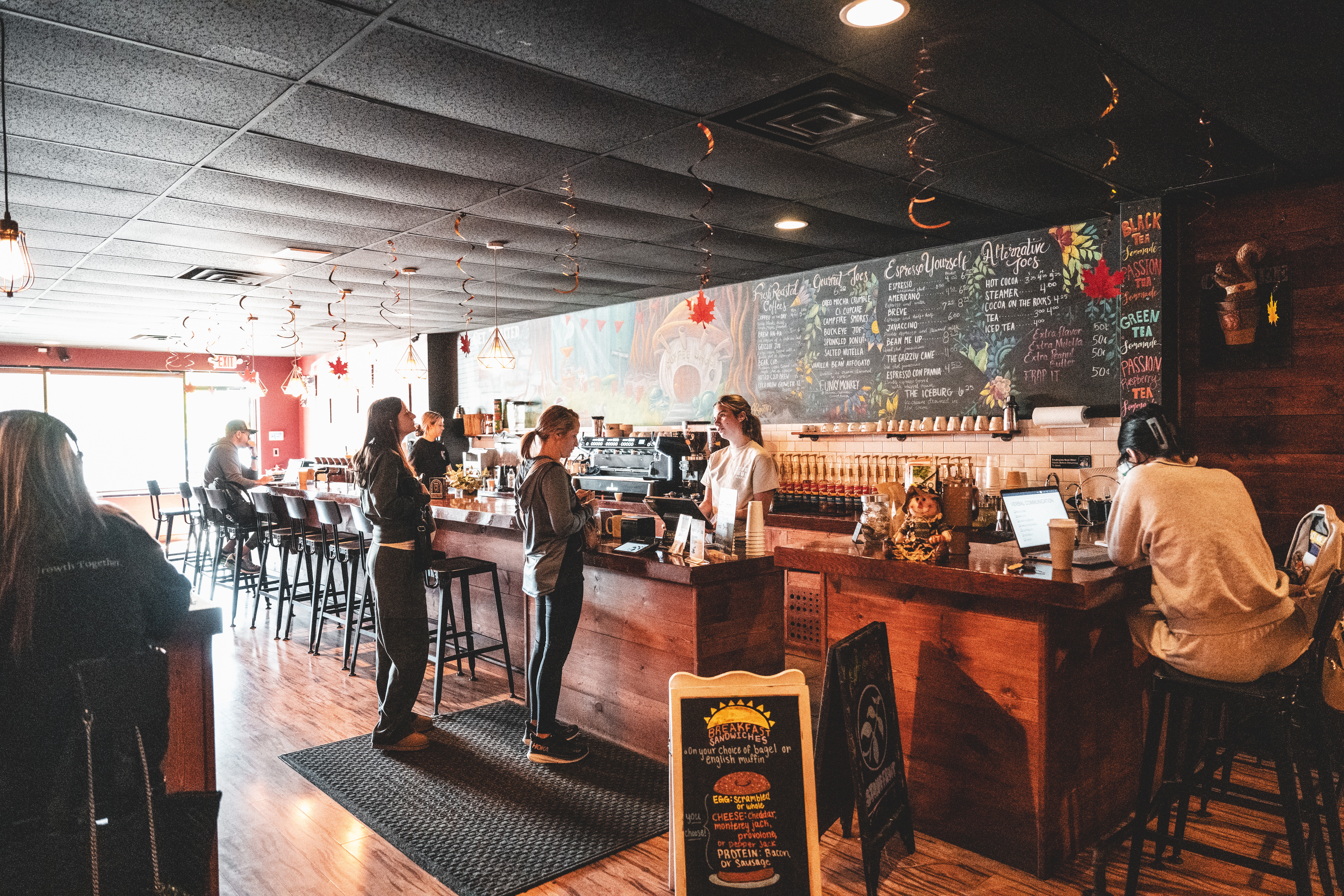People standing at a bar counter in a restaurant with a chalkboard menu on the wall, dim lighting, and seating area.