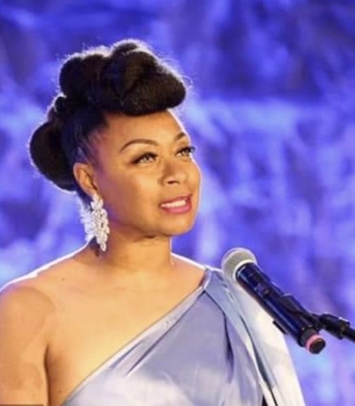 Woman with styled hair and earrings speaking into a microphone against a blue background.