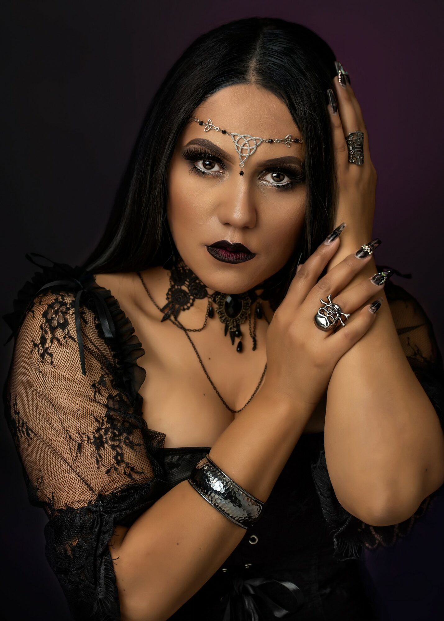 Woman with dark makeup and jewelry, wearing black lace clothing, posing with hands near face against dark background.