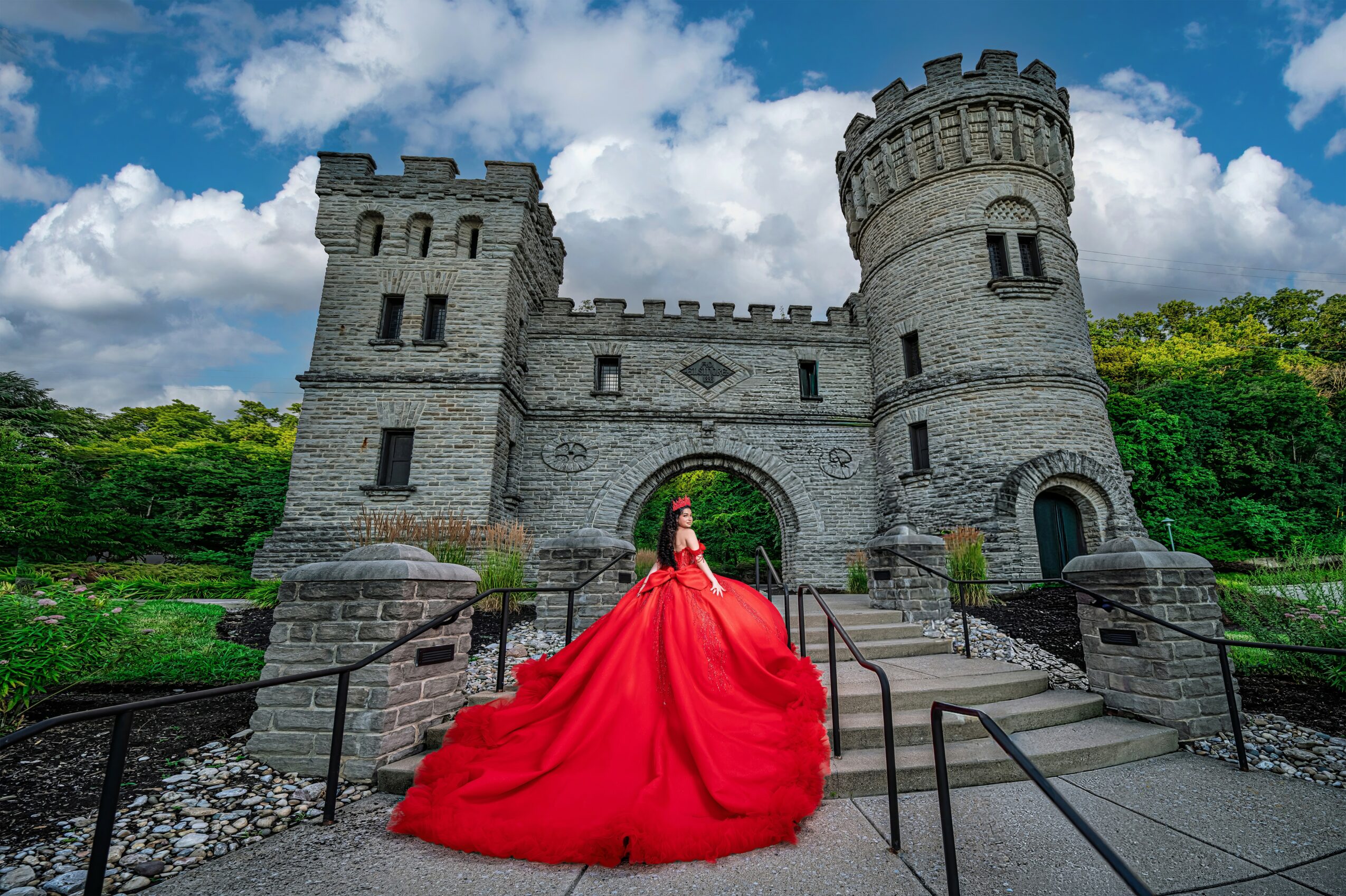 Person in a red dress standing on a stone bridge in front of a castle with towers, under a partly cloudy sky.