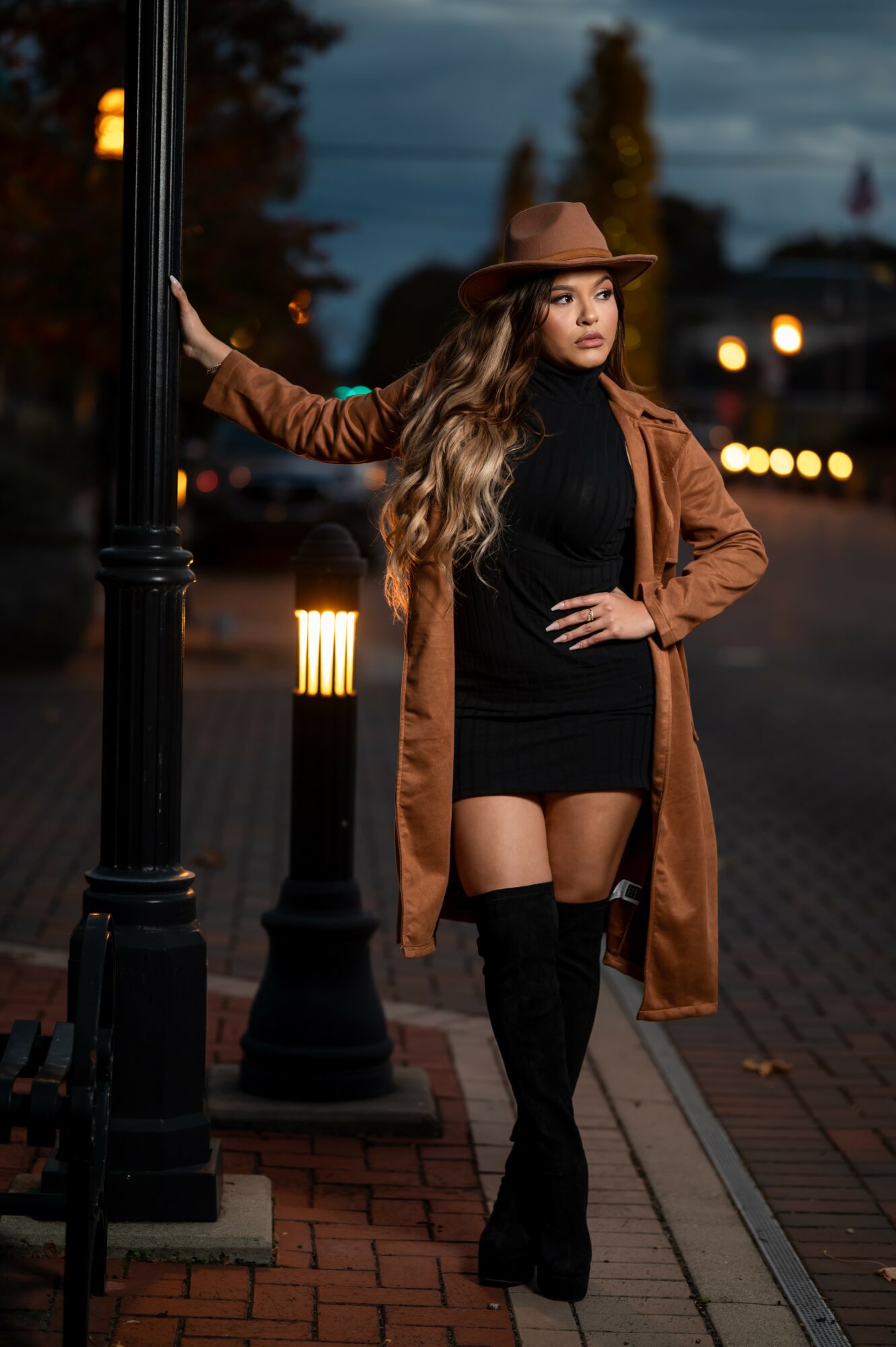 Woman in black outfit and brown coat with hat standing on sidewalk at dusk, holding a pole, streetlights in background.