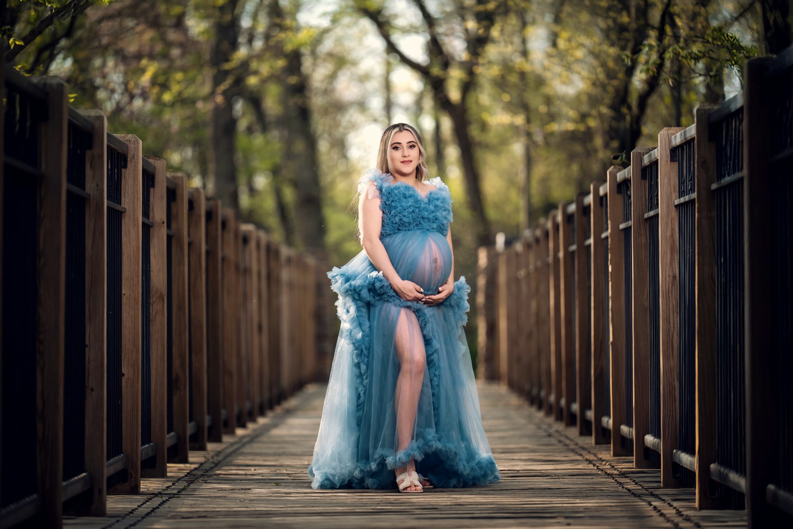 Woman in a blue dress holding a pregnant belly on a wooden bridge, trees in background.