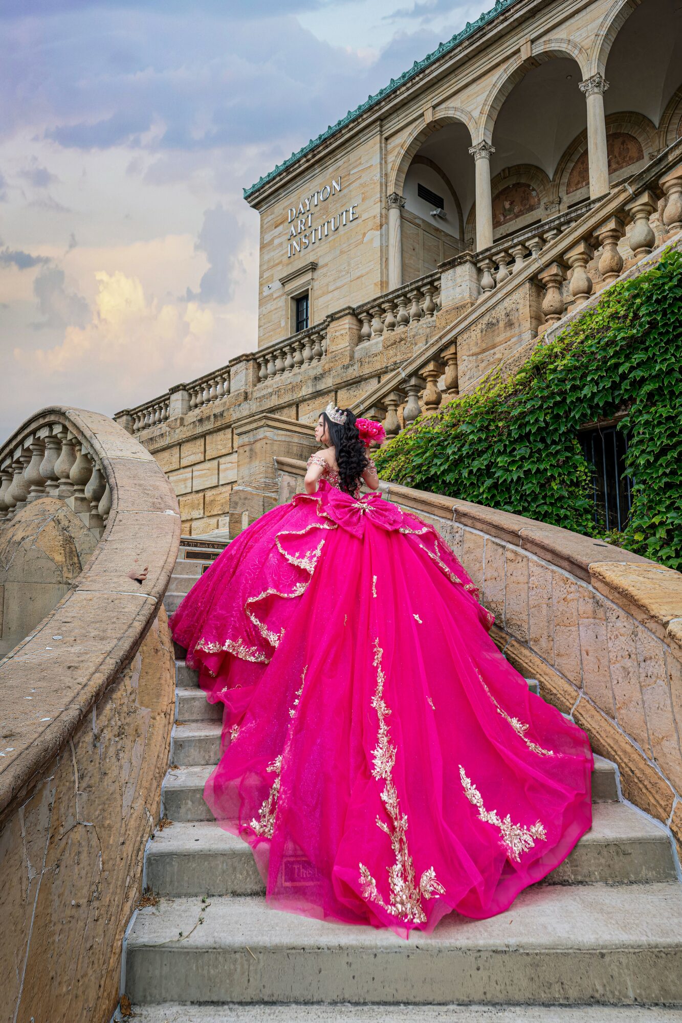 Person in a bright pink gown with gold embroidery ascending stone stairs outside a historic building with arches and greenery
