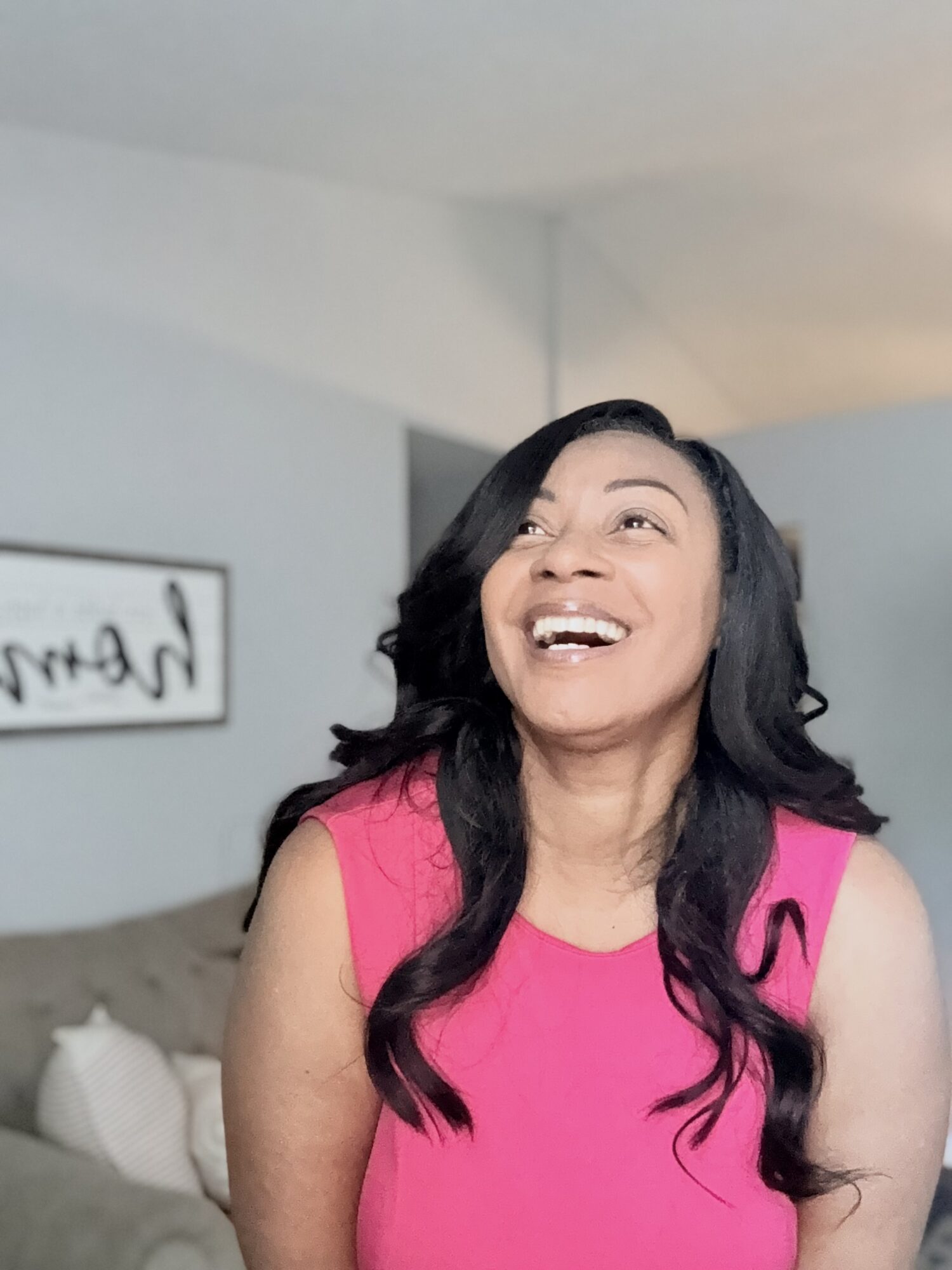 Woman with long black hair smiling, wearing a pink sleeveless top, in a room with a gray wall and a framed picture.