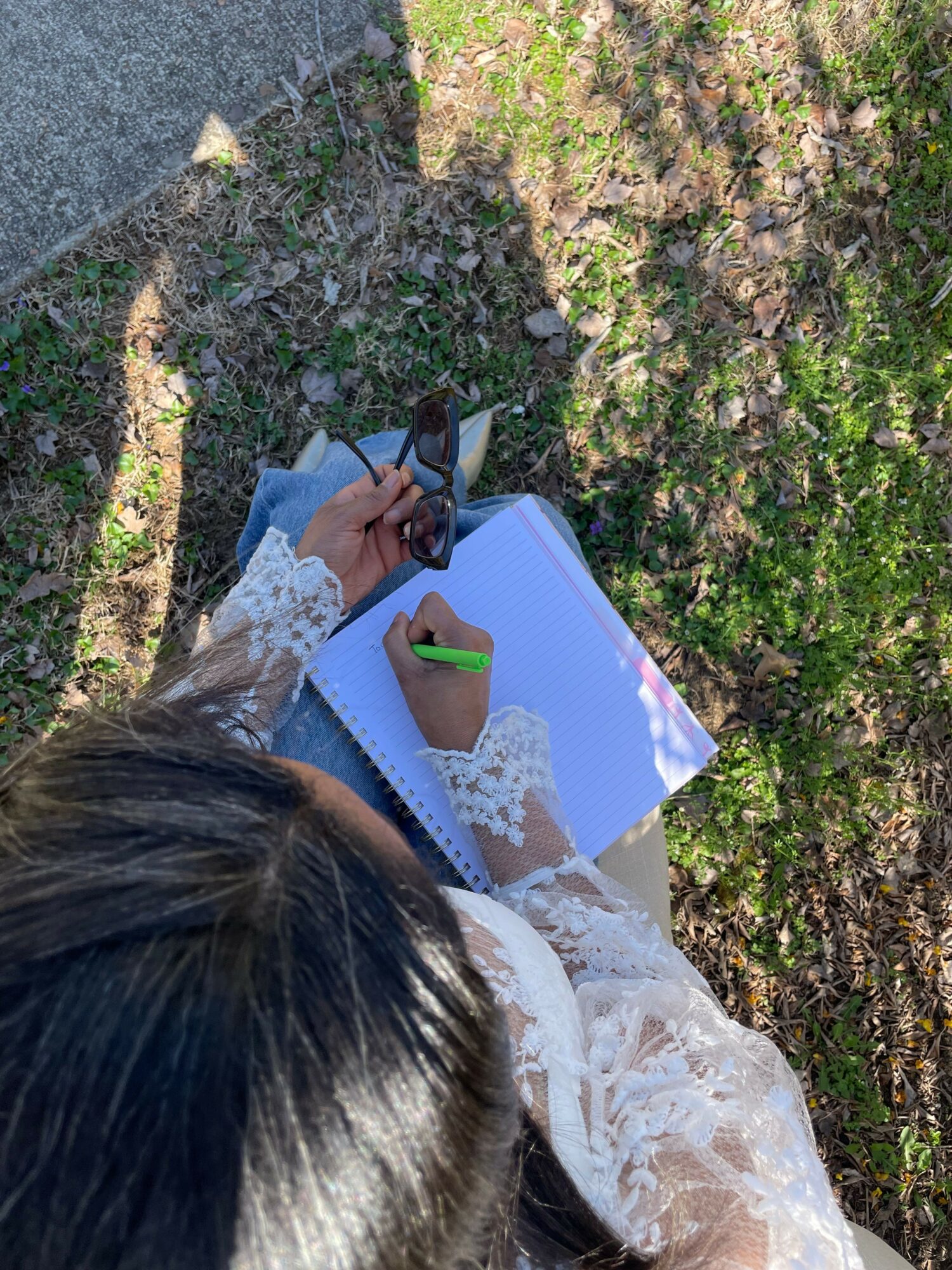 Person with long dark hair writing in a notebook outdoors, with a green pen, on grass and dirt.