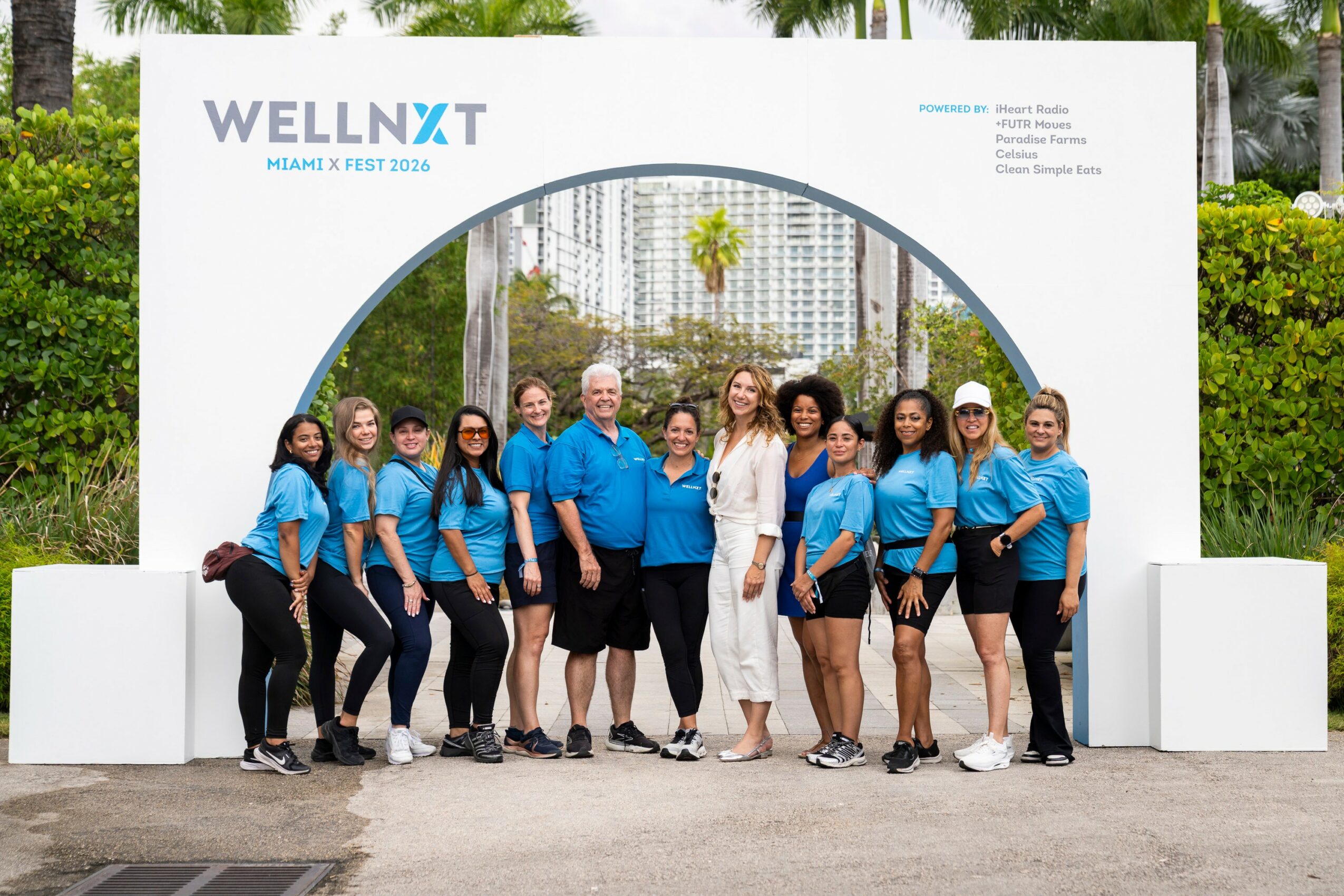 Group of people in blue shirts standing outdoors in front of a large white display with a circular opening, cityscape background.