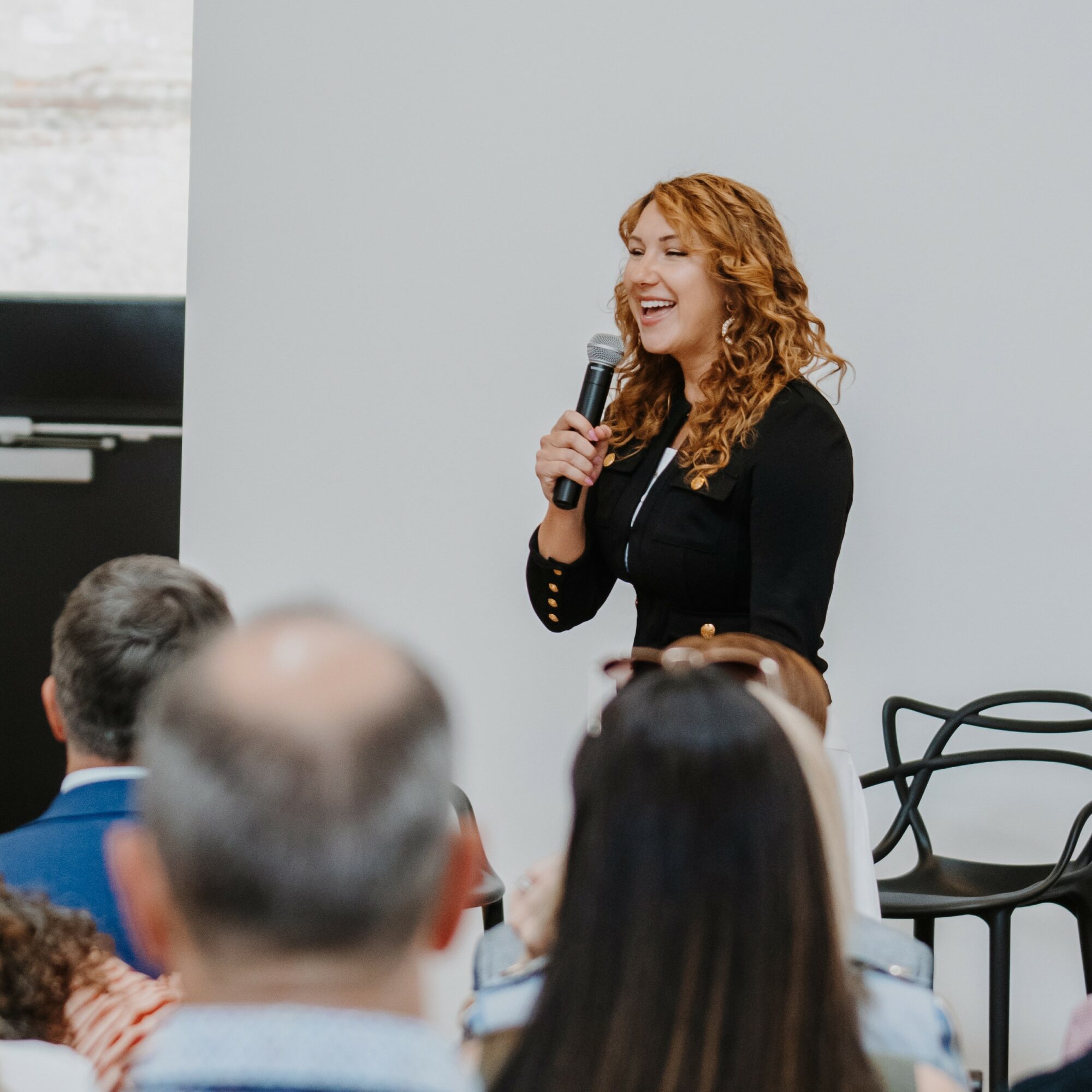 Woman with curly hair speaking into a microphone in front of an audience, smiling, with a plain background.