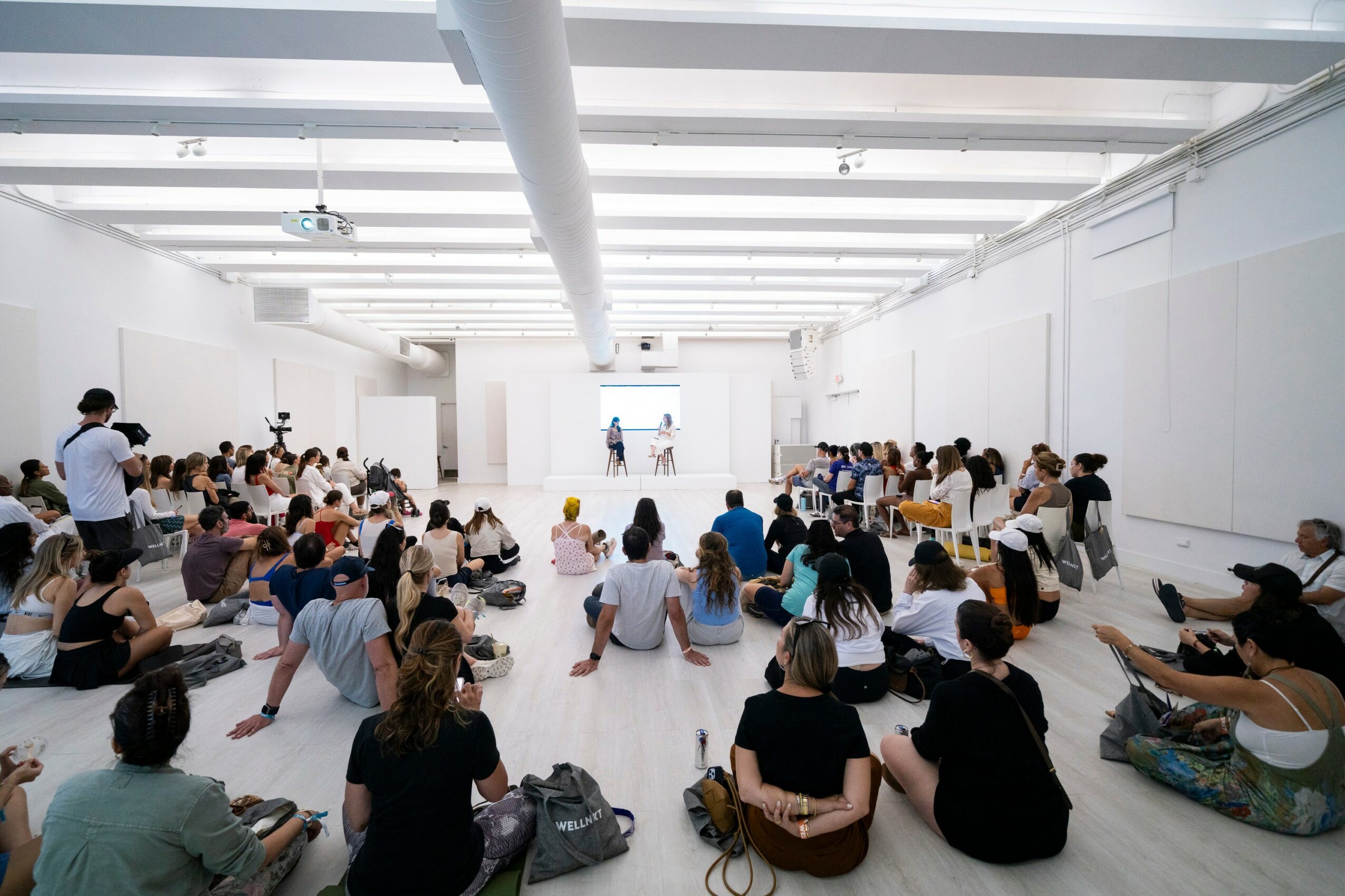 Large group of people sitting on the floor in a spacious, white-walled room, facing a stage with a person speaking.