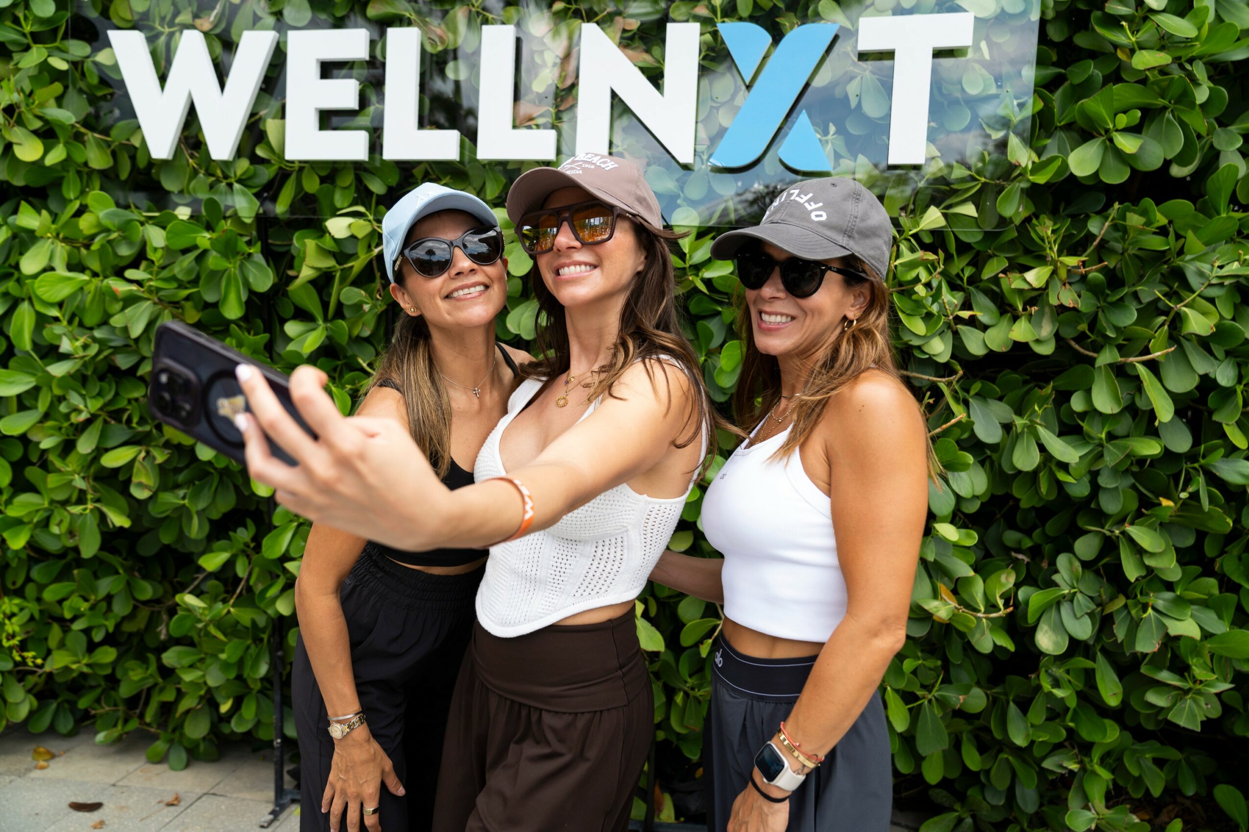 Three women taking a selfie in front of a green leafy background with a sign that reads 'WELLNXT'.