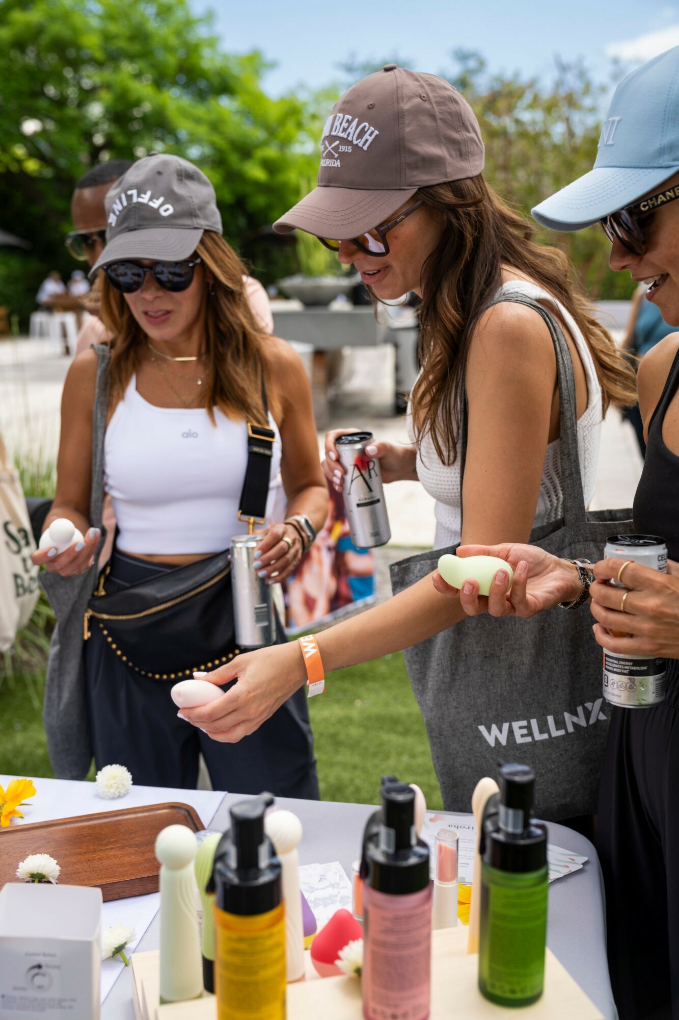 Three women outdoors at a table with skincare products, holding a hot dog, wearing hats and sunglasses.