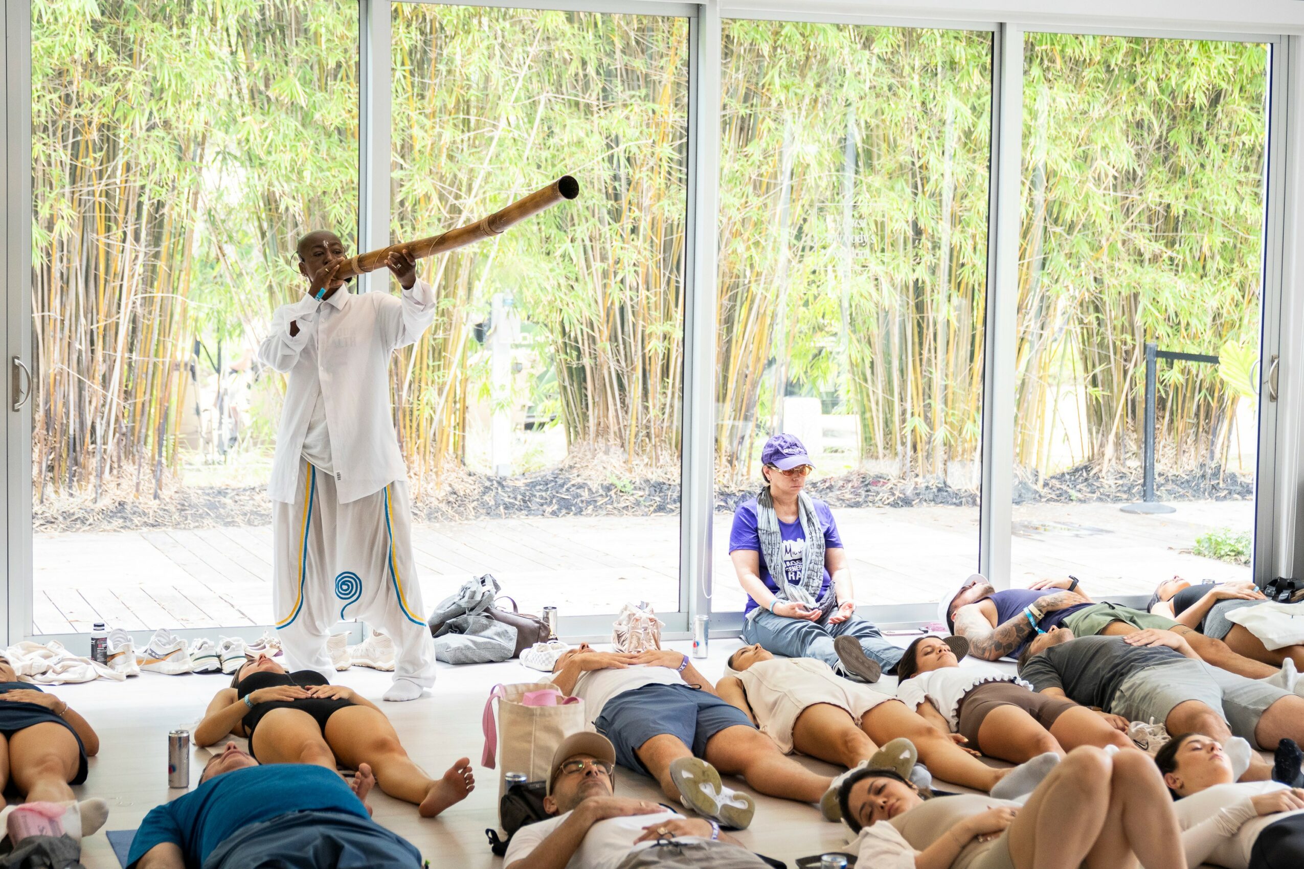 Person standing and playing a traditional instrument, audience lying on the floor, large windows with trees outside, bright room.