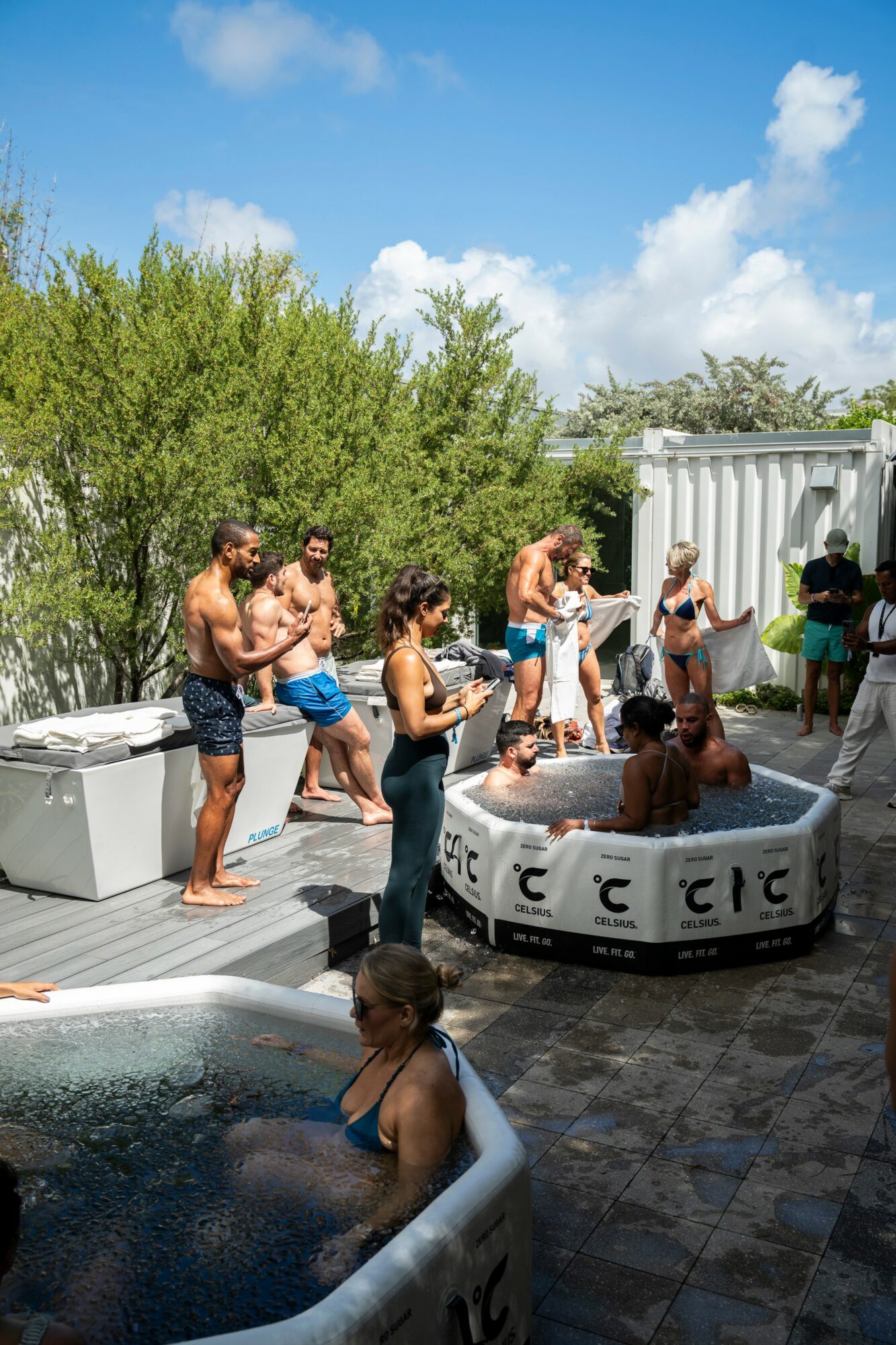 People enjoying a pool party outdoors with a hot tub, trees, and a blue sky with clouds in the background.