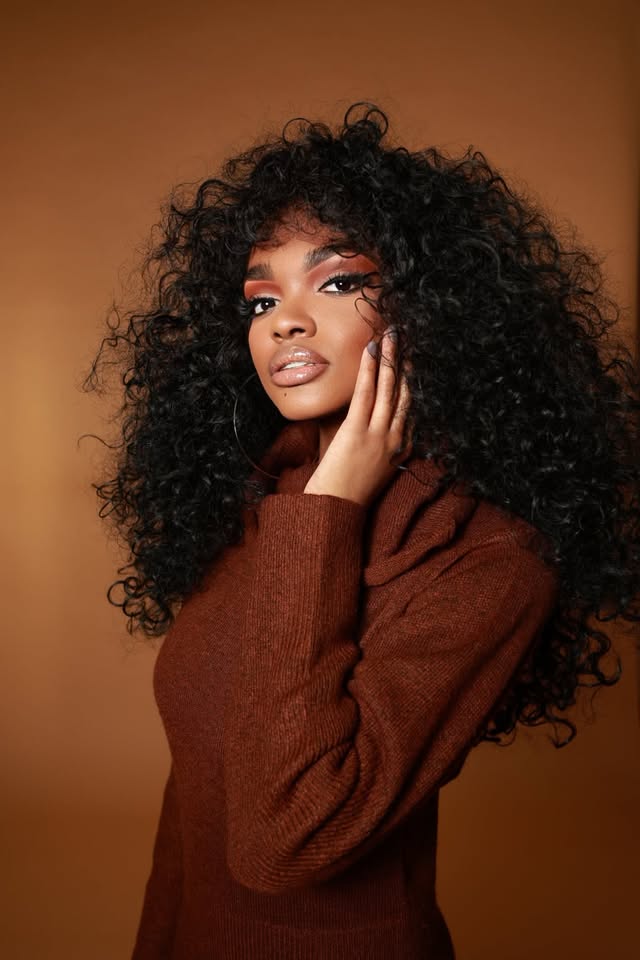 Young woman with voluminous curly hair, wearing a brown sweater, posing with hand near face against a warm background.