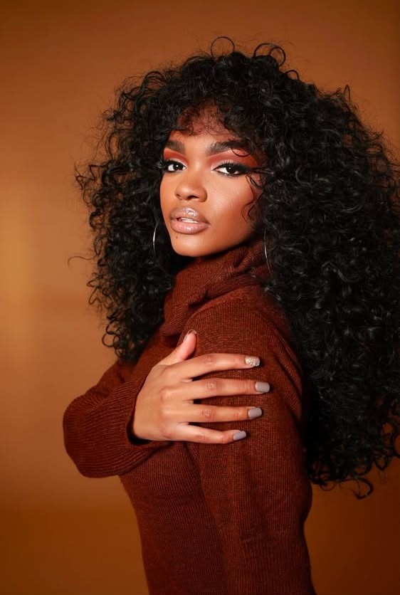 Young woman with curly hair and makeup, wearing a brown sweater, looking at the camera, warm background.
