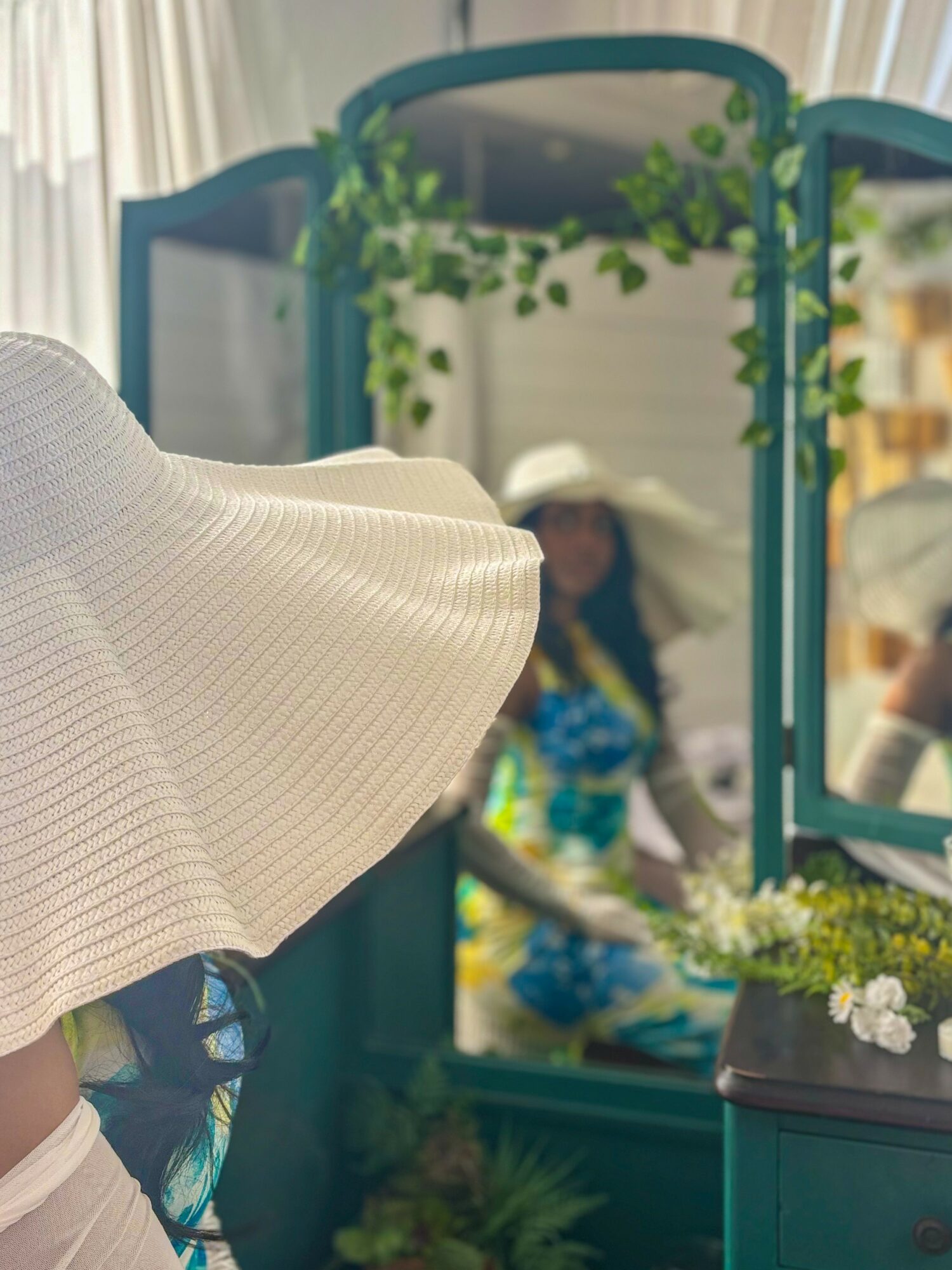 Person in white clothing looking at a mirror with a woman in colorful dress reflected, decorated with greenery and flowers.