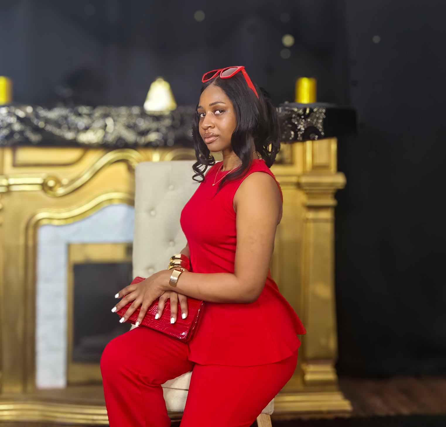 Woman in red outfit sitting on a chair, holding a red clutch, with a gold and black background.
