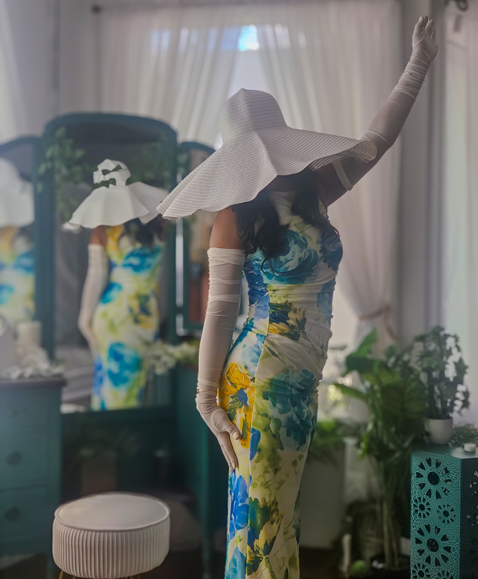 Person in a floral dress with a large white hat and gloves, standing in a decorated room with plants and curtains.