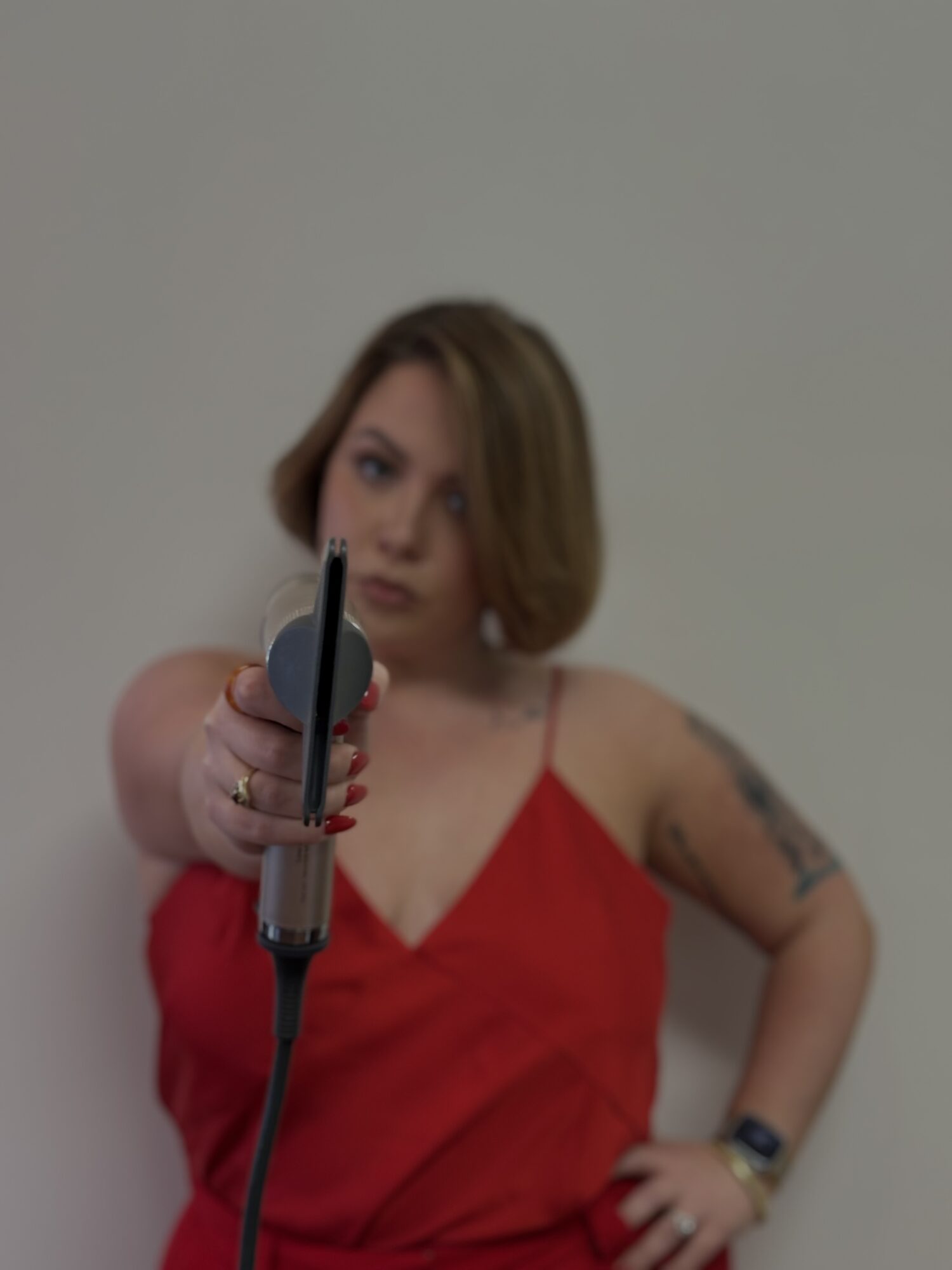 Woman in red dress pointing a gun at the camera, with tattoos on her arm, against a plain background.