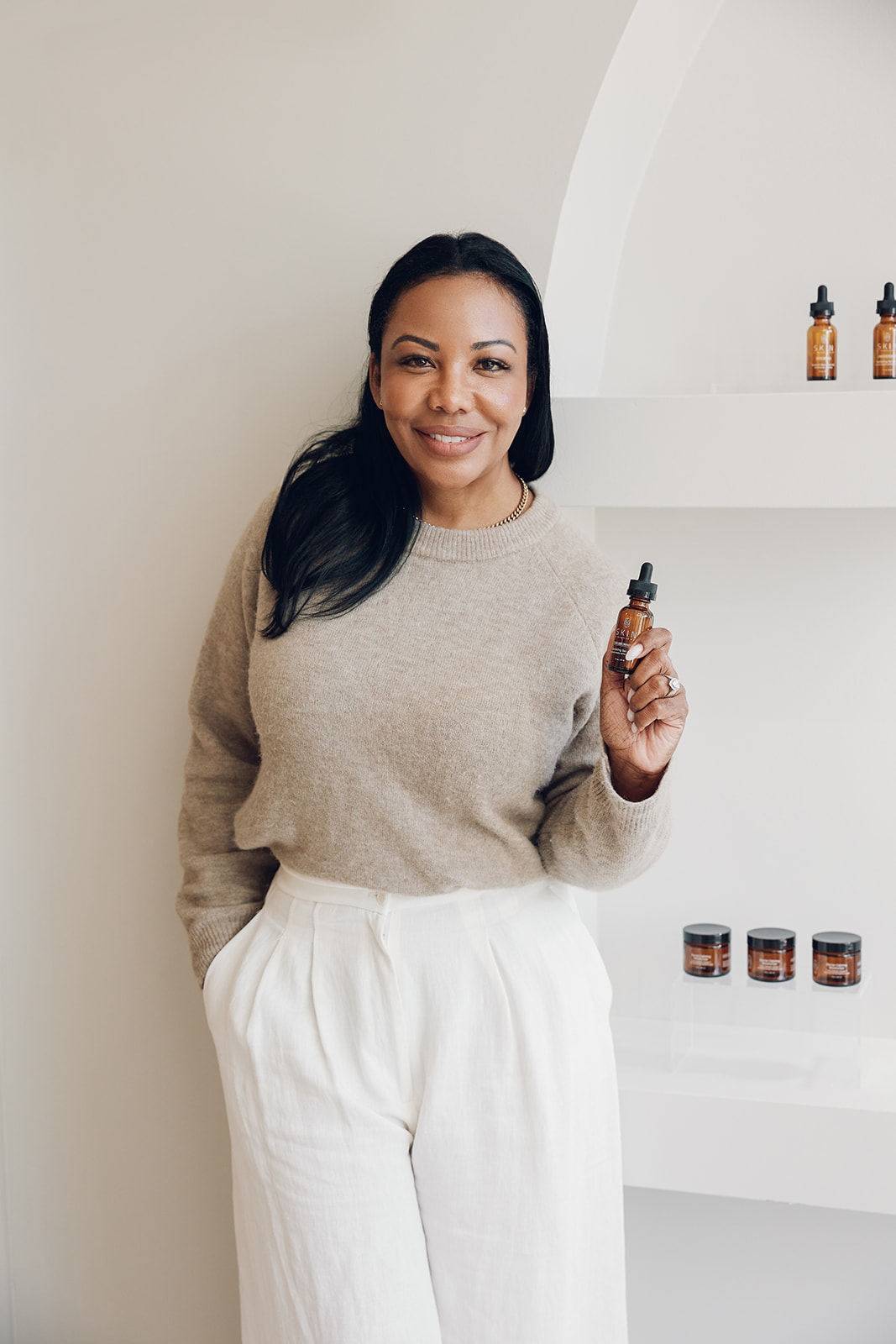 Woman with long dark hair smiling, holding a small bottle, standing in front of a white wall with shelves and bottles.