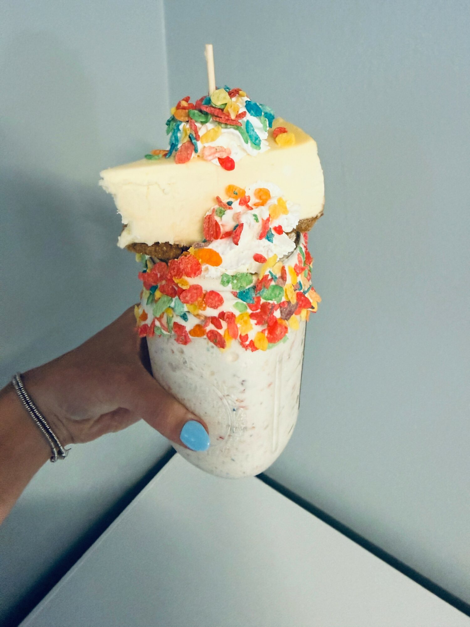 Hand holding a colorful ice cream float with sprinkles and cake on top, against a light blue wall.