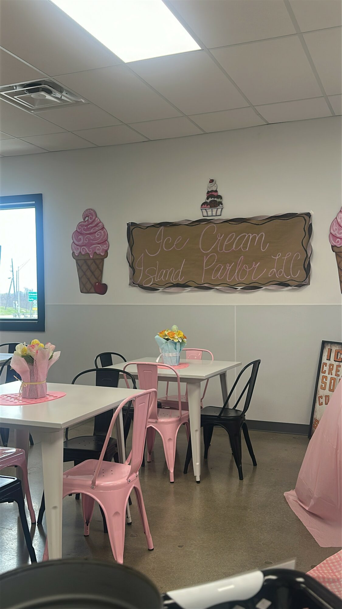 Indoor space with tables, chairs, and wall decorations including ice cream cone cutouts and a sign, with a window on the left.