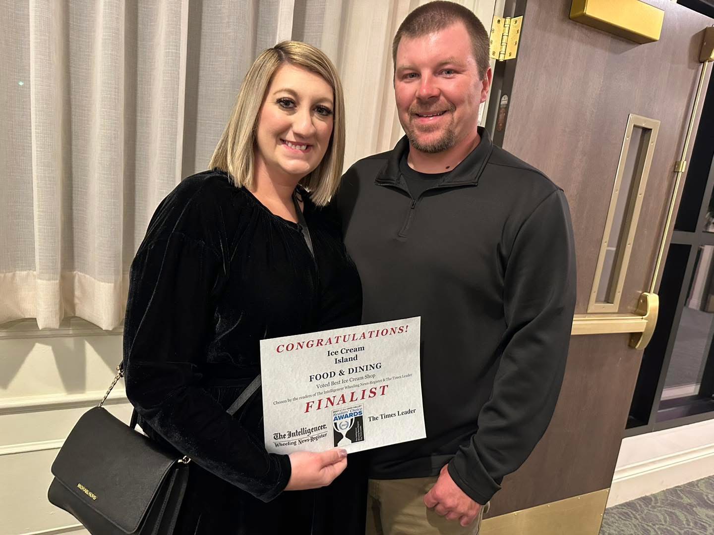 A woman and a man holding a certificate, smiling indoors with curtains and a wall behind them.