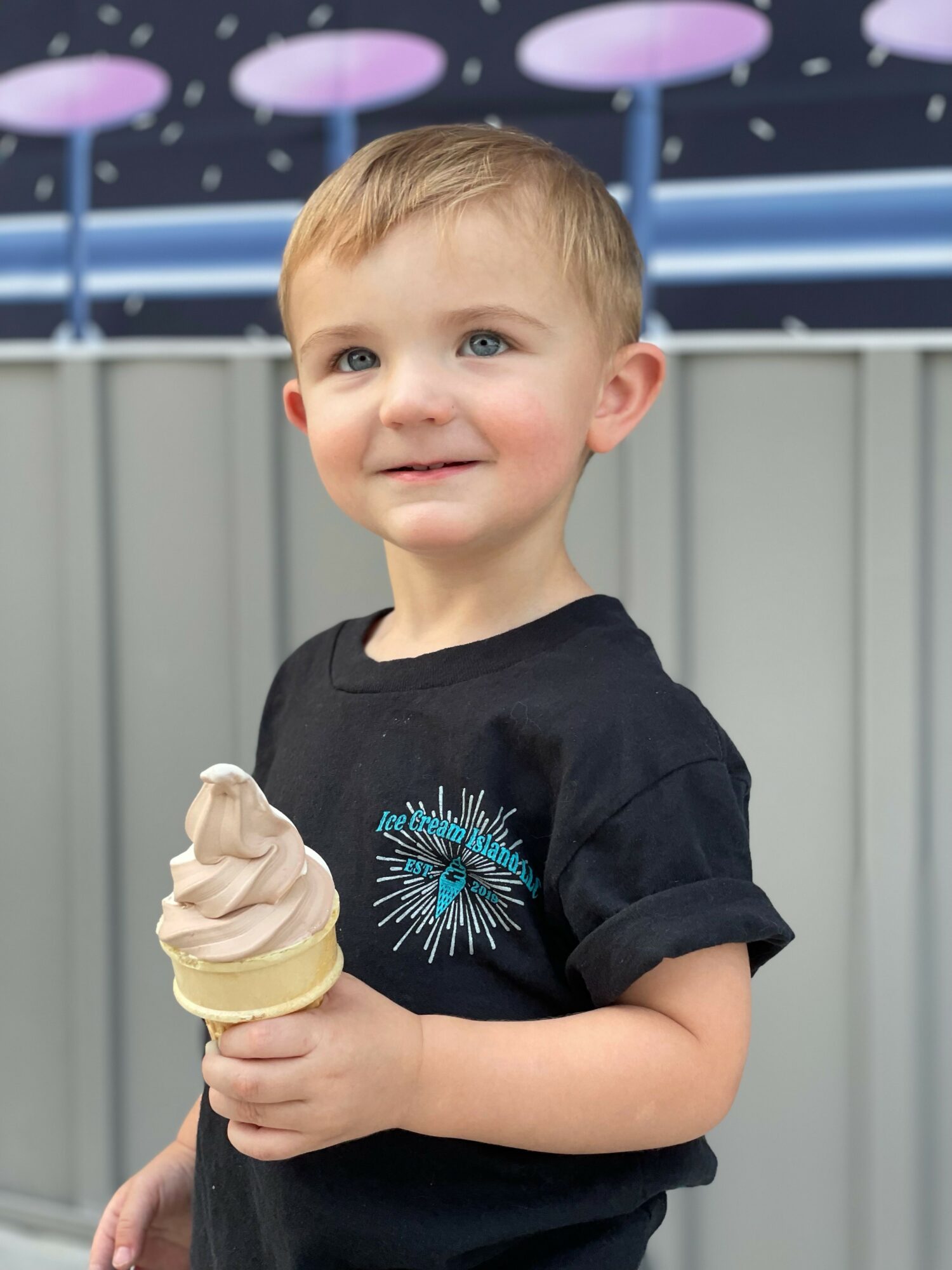 Young boy holding ice cream cone, smiling outdoors, gray fence and purple umbrellas in background.