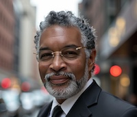 A smiling man with gray hair and beard, wearing glasses and a suit, standing outdoors in an urban setting.