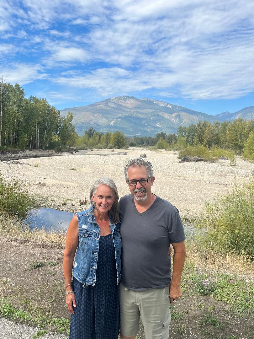 A smiling woman and man stand outdoors with mountains, trees, and a river in the background under a partly cloudy sky.