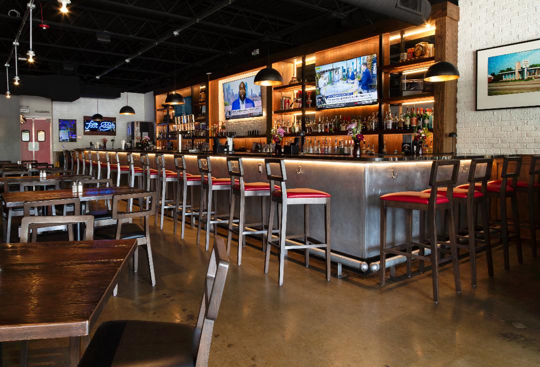 Bar with high stools, multiple TV screens, and shelves with bottles, in a modern restaurant setting.