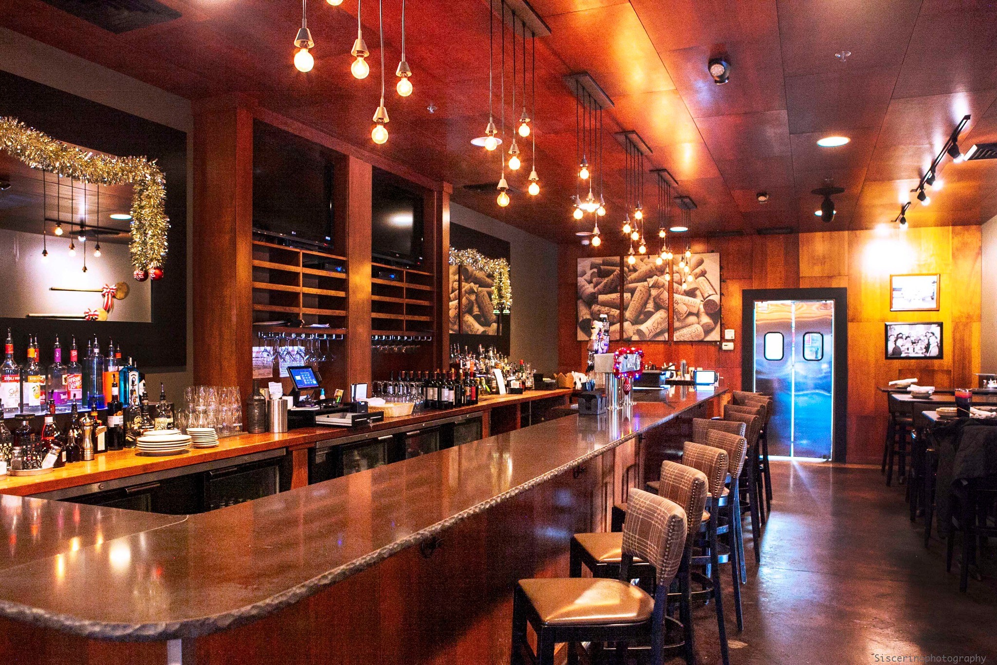 Bar interior with wooden counter, hanging lights, shelves with bottles, and seating area, decorated for the holidays.
