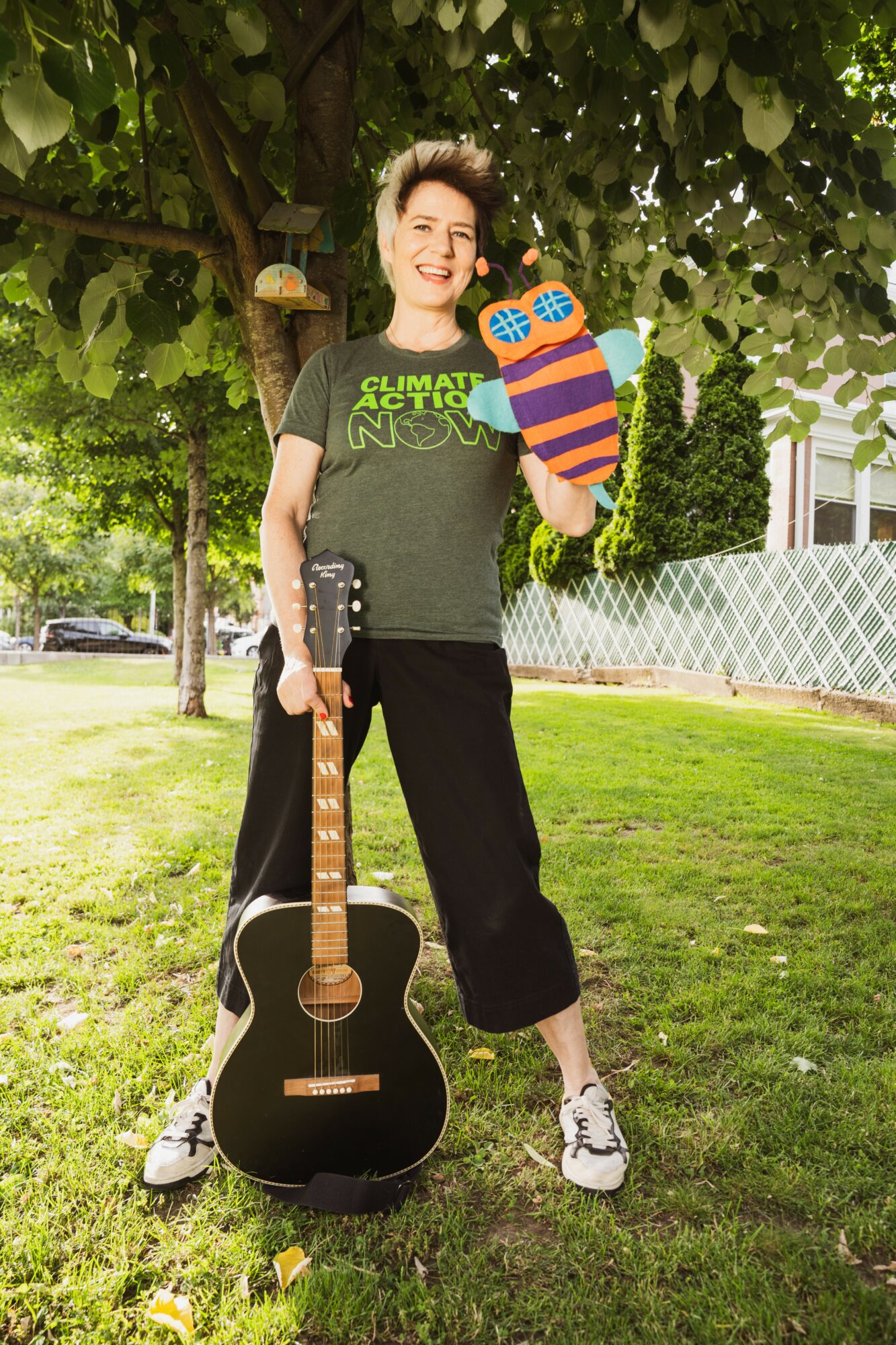 Person standing outdoors under a tree, holding a guitar and a colorful sock puppet, smiling, with a grassy yard and fence in background.