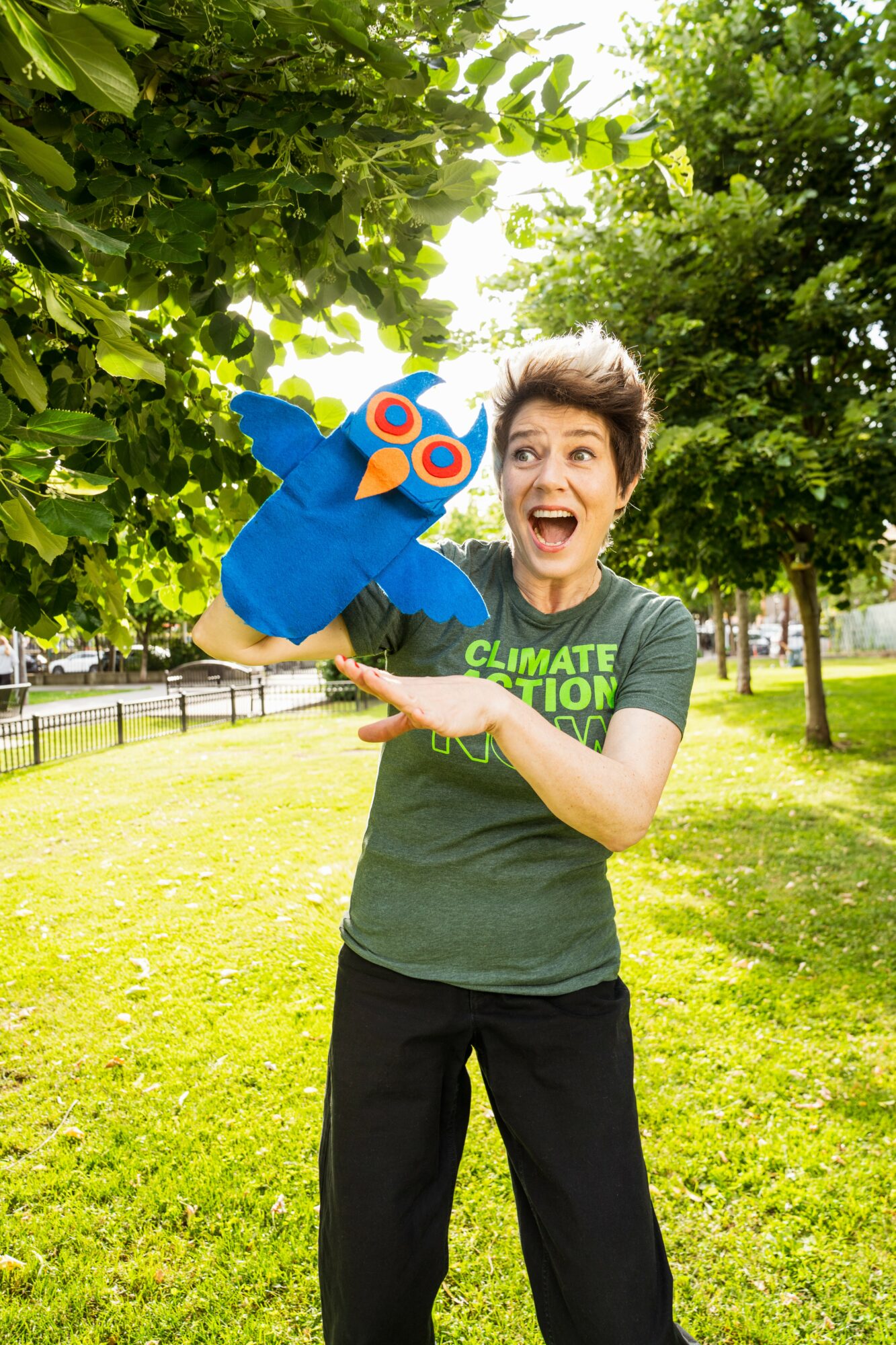 Person holding a blue puppet with orange eyes outdoors, smiling, surrounded by green trees and grass.