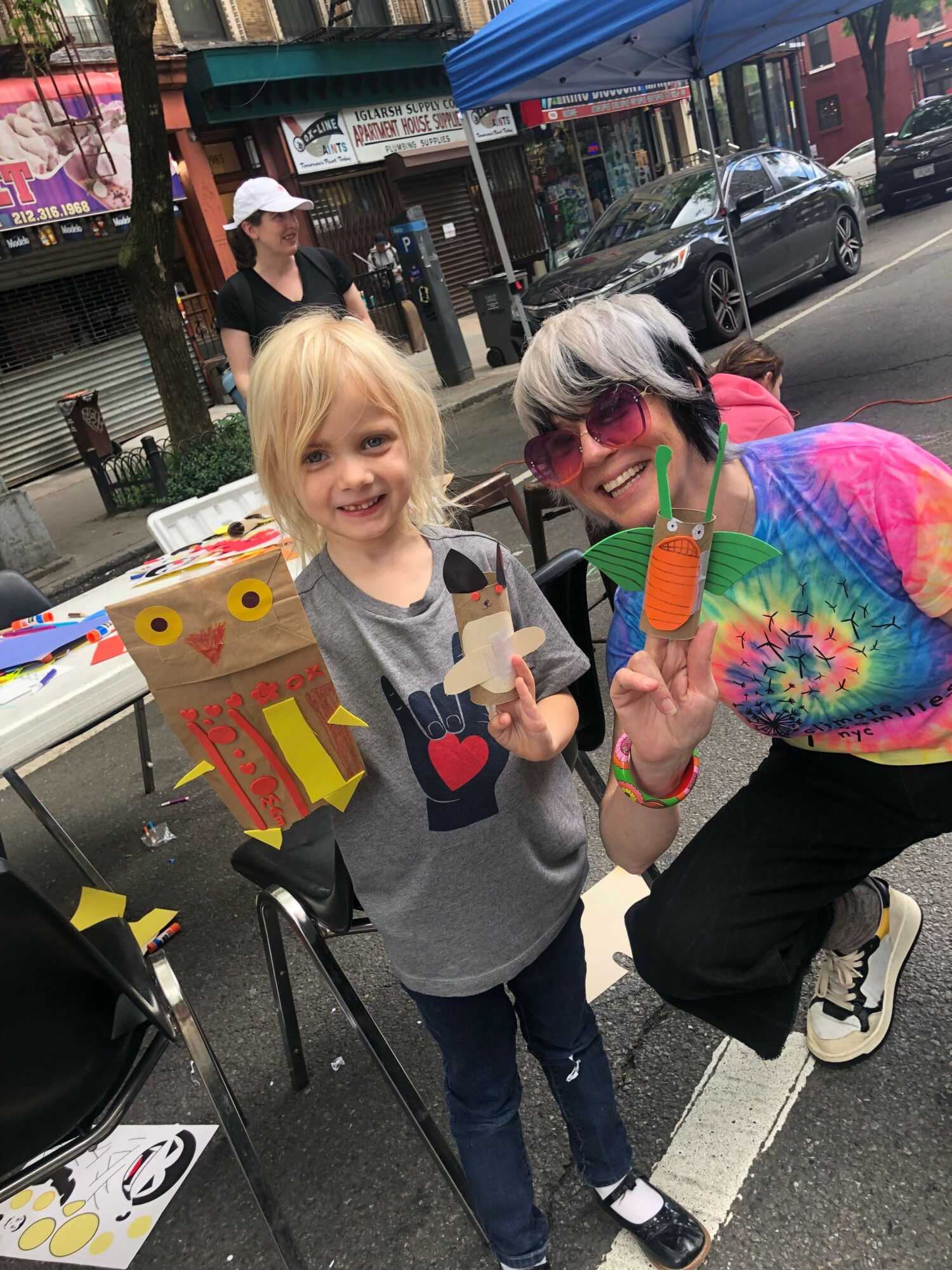 Two people standing outdoors near a table with colorful decorations, one holding a star-shaped object, the other making a peace sign.