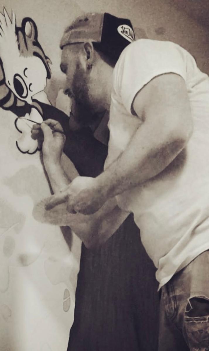 Person drawing on a wall with cartoon character, wearing a cap and casual clothes, leaning close to the wall.