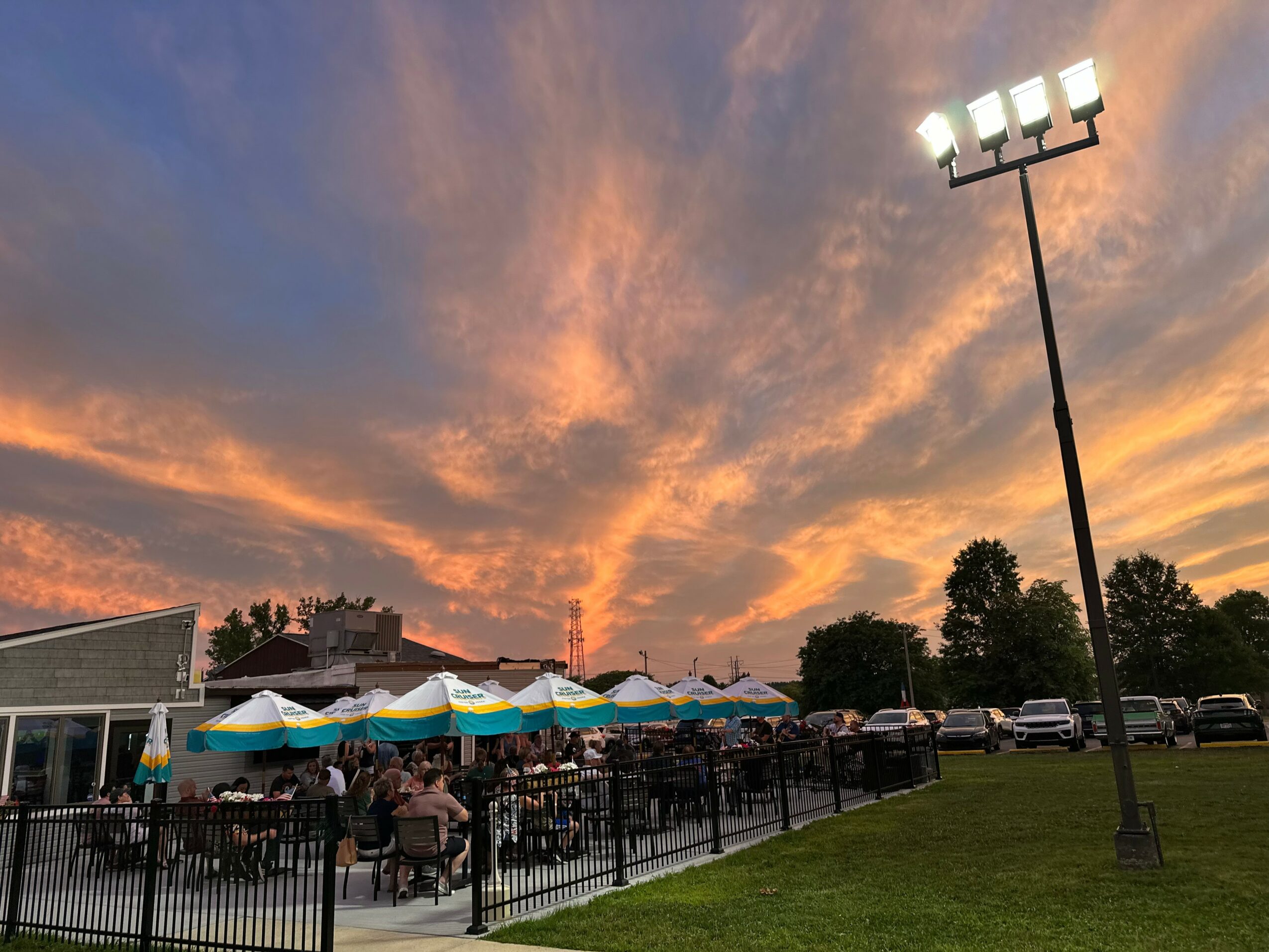 Sunset sky with orange and blue clouds over outdoor seating area with umbrellas, people, and parked cars, illuminated by tall floodlight.