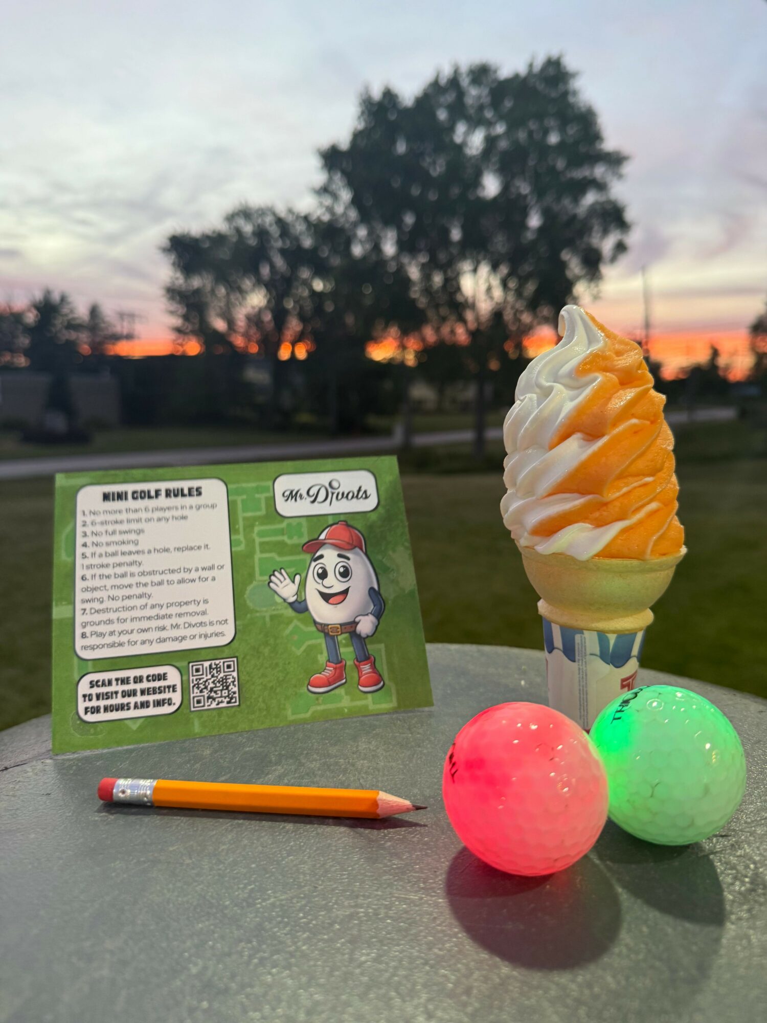 Ice cream cone with orange and white swirl, two colorful balls, a pen, and a card on a gray surface outdoors at sunset.