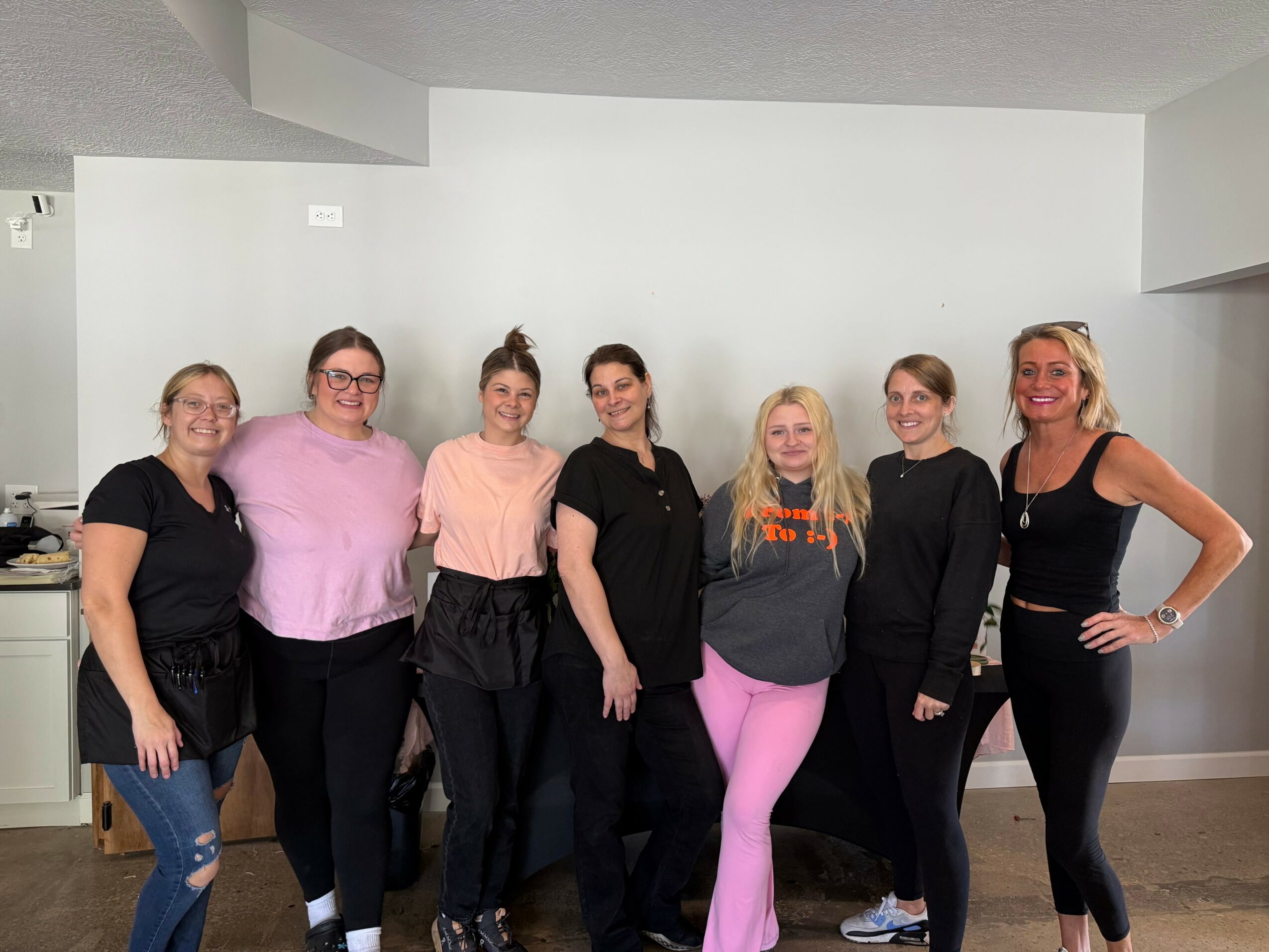 Seven women standing in a row indoors, smiling, with a plain white wall background.