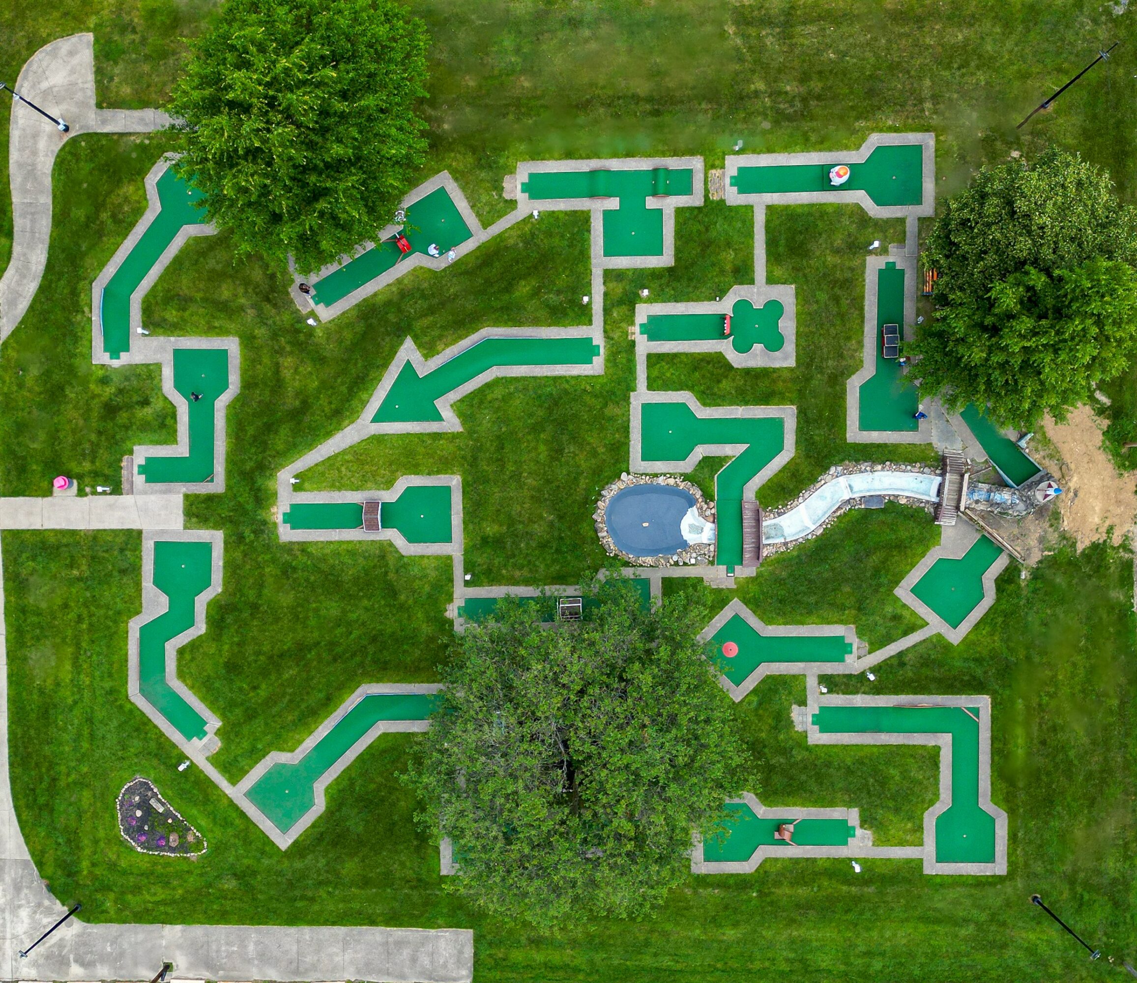 A playground with multiple interconnected structures, slides, and a central circular area with a blue surface, surrounded by green grass.