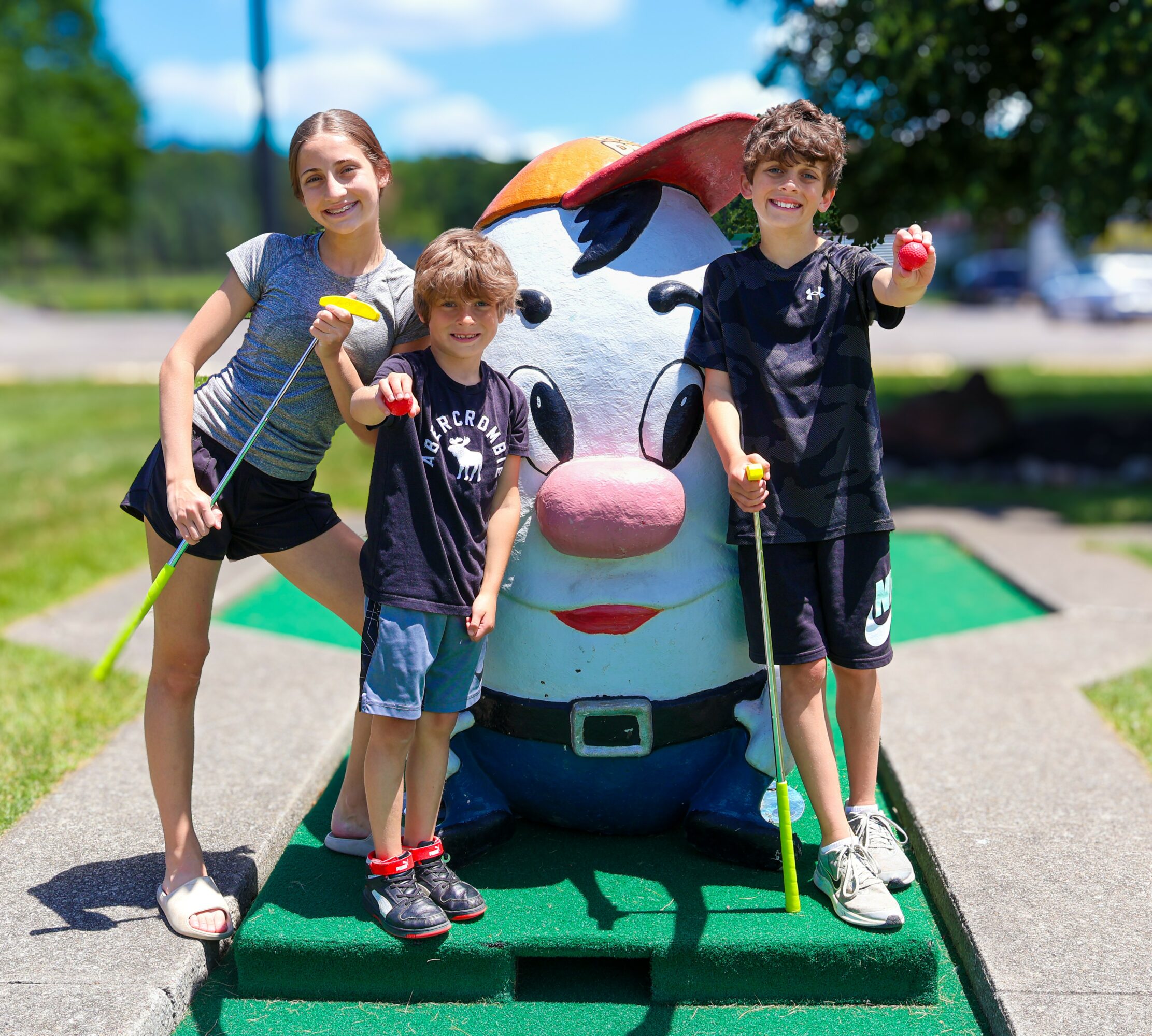 Three children with a large cartoon dog statue outdoors on a sunny day, smiling and holding mini golf clubs.