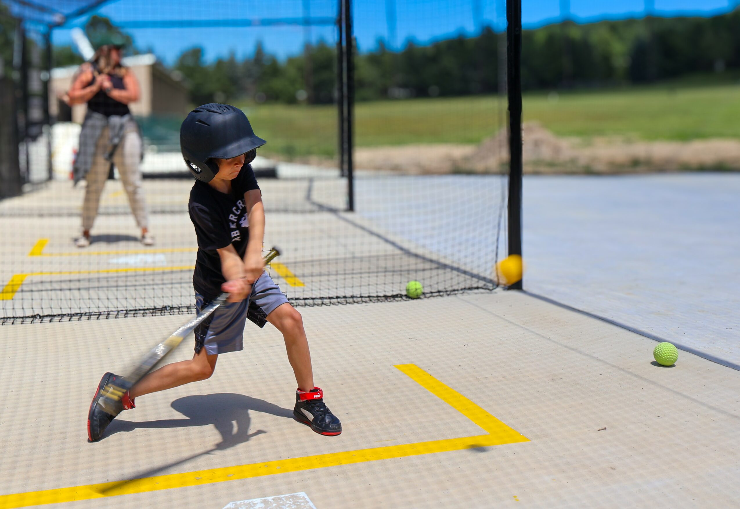 Child in black shirt and helmet swings a baseball bat at a tennis ball on a court, with a woman watching in the background.