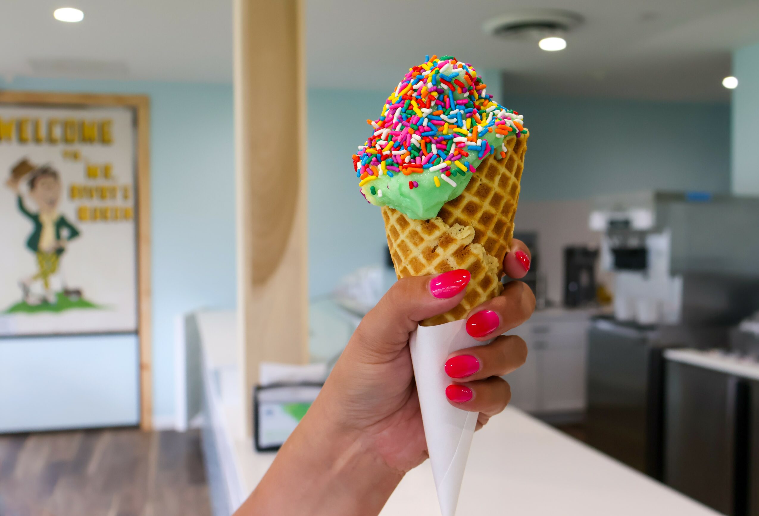 Hand holding a colorful ice cream cone with sprinkles inside a shop, background includes a welcome sign and kitchen area.